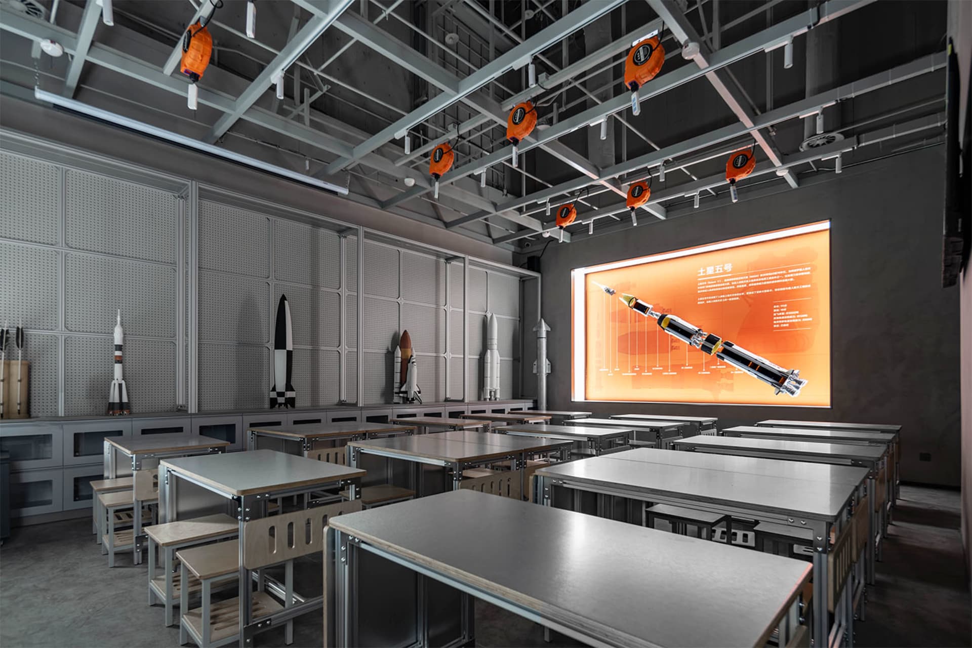 Modern STEM classroom featuring rows of metal and wood desks, rocket and spacecraft models, large digital screen displaying a rocket diagram, and bright orange industrial pendant lights hanging from an exposed ceiling.