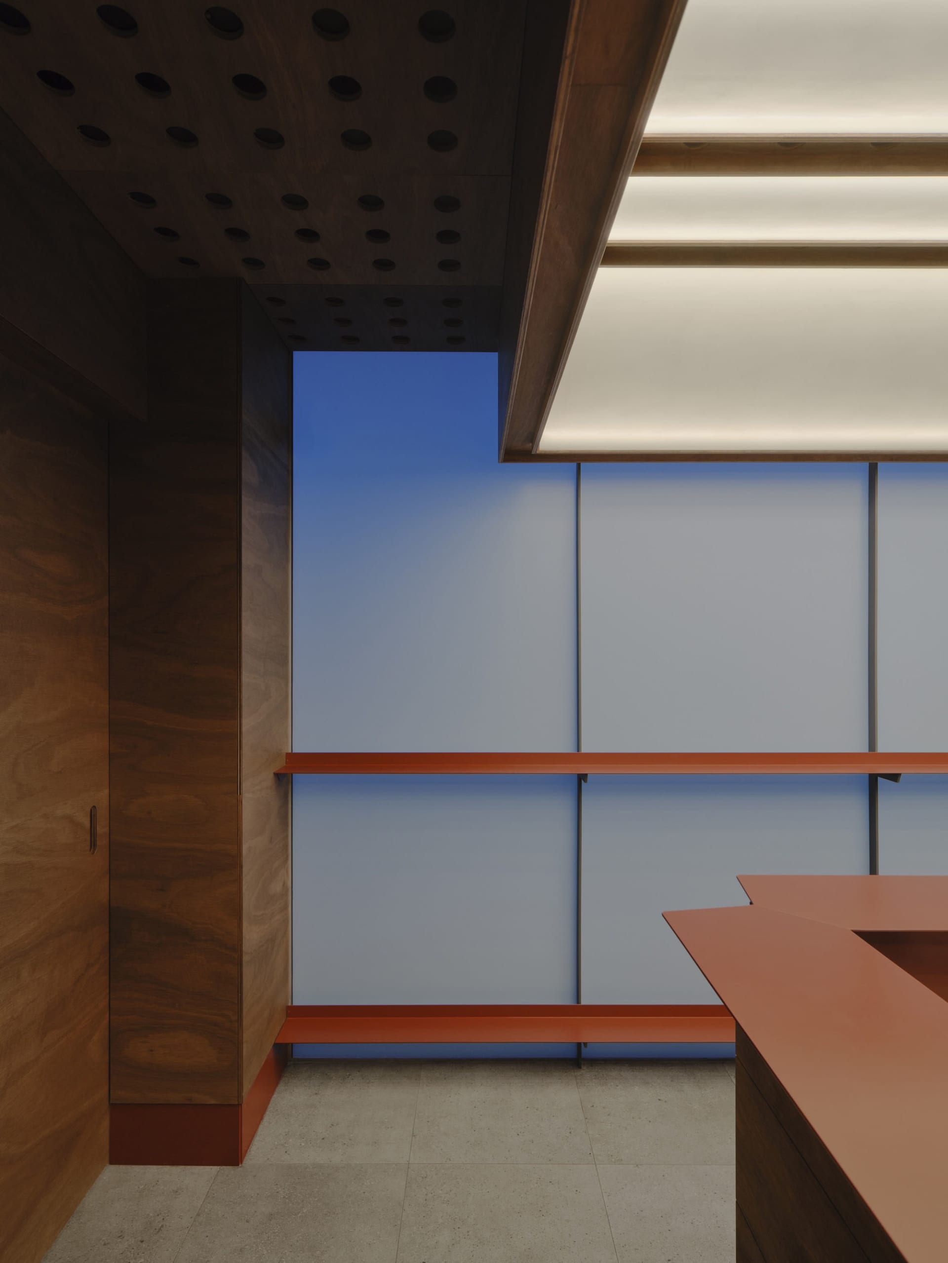 Modern reception area featuring dark walnut wood paneling, a terracotta-colored desk, concrete floor, and a large blue backlit wall panel under a wooden ceiling with circular acoustic perforations.