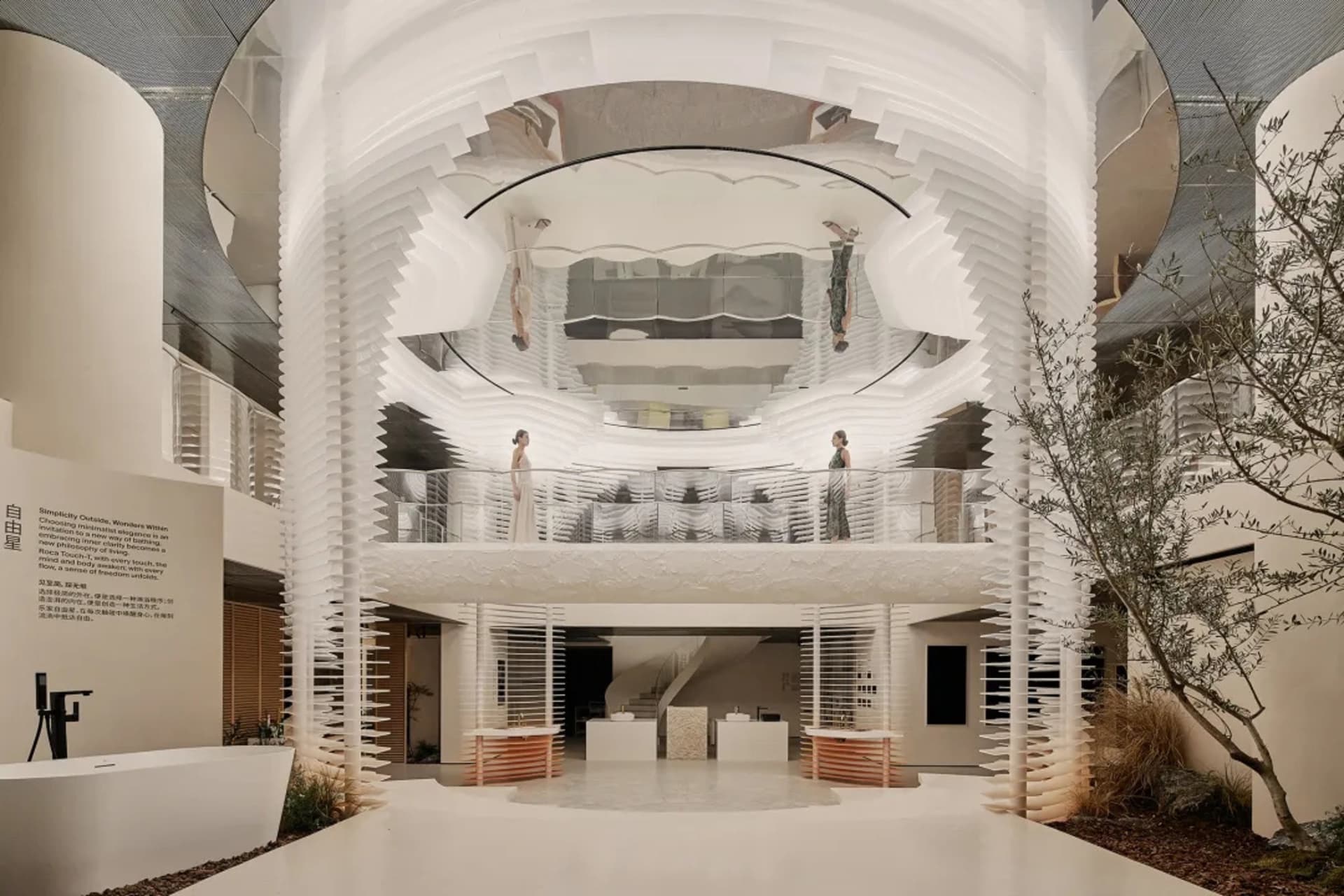 Modern multi-story white atrium interior with layered 3D textured walls, glass railings, olive tree, and people standing on balconies.
