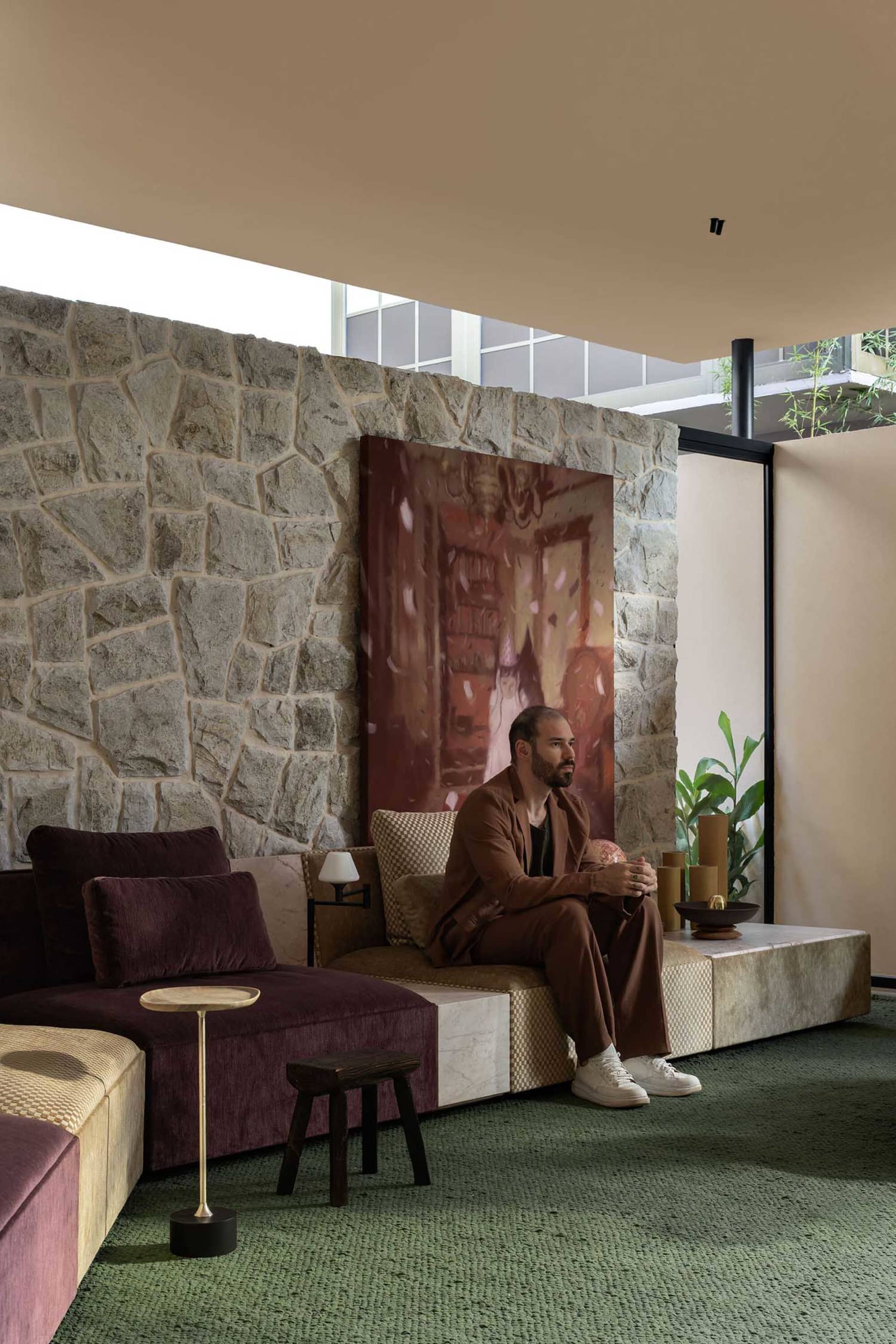 Man in brown suit sitting on a modular sofa with maroon velvet and checkered upholstery in front of a large natural stone wall featuring a dramatic painting, green carpeted floor, and indoor plants.