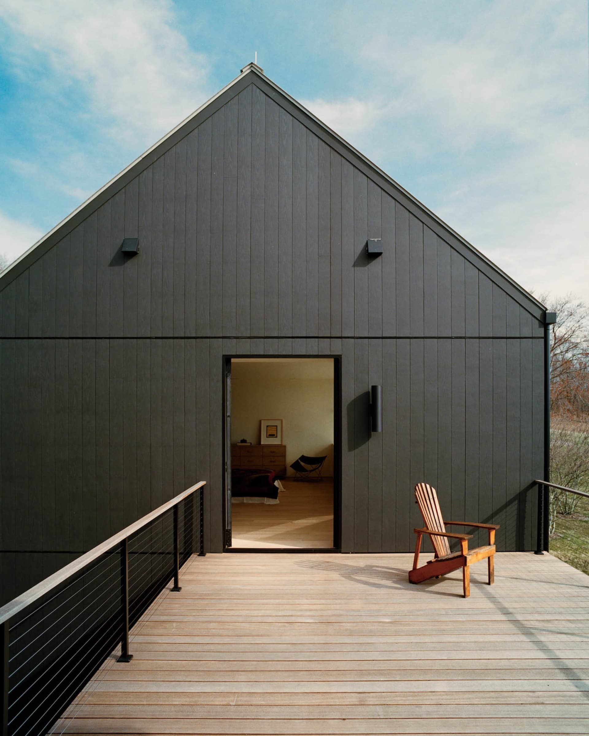 Modern black barn house with large gable roof, open sliding door revealing a bedroom interior, wooden deck with railing, and a single red Adirondack chair under a blue sky.