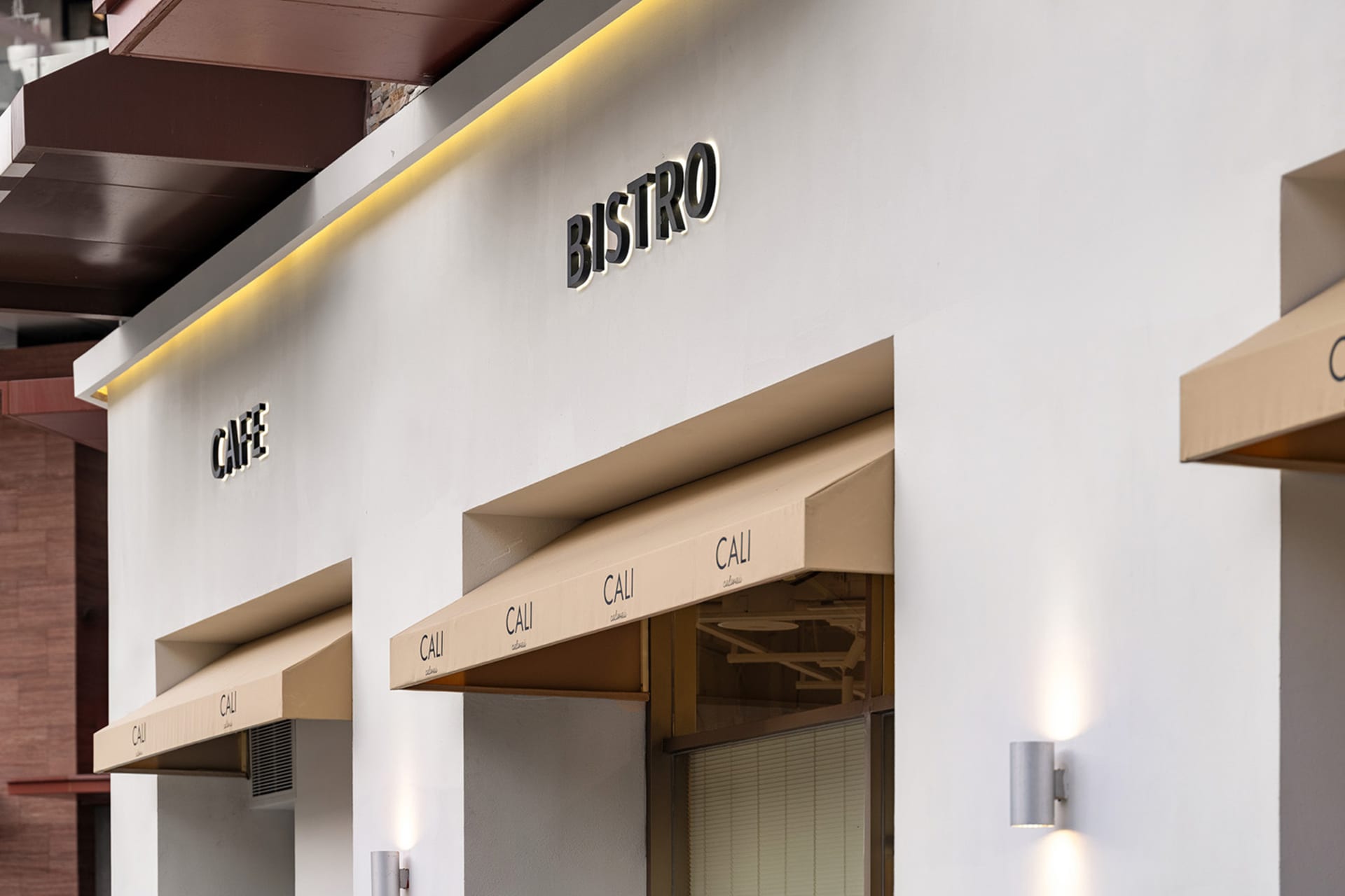 Modern white commercial facade of a cafe and bistro with beige fabric awnings, illuminated 'CAFE' and 'BISTRO' signs, and cylindrical wall sconces.