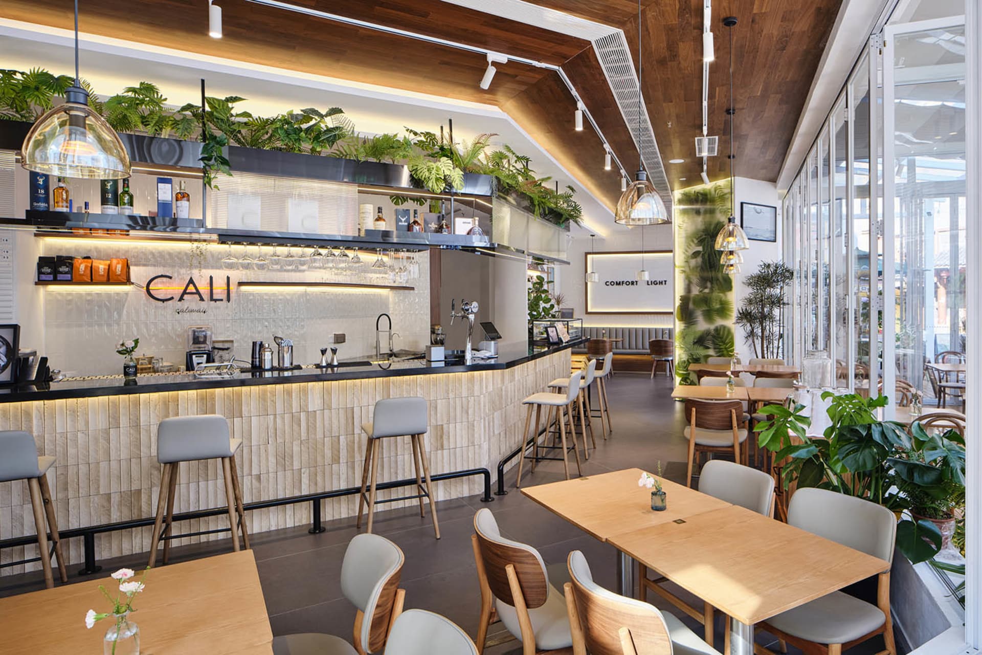 Bright modern cafe interior with a long wooden bar counter, grey upholstered bar stools, abundant hanging ferns and monstera plants, light wood dining tables and matching chairs, warm wood ceiling, and large glass windows allowing natural light.