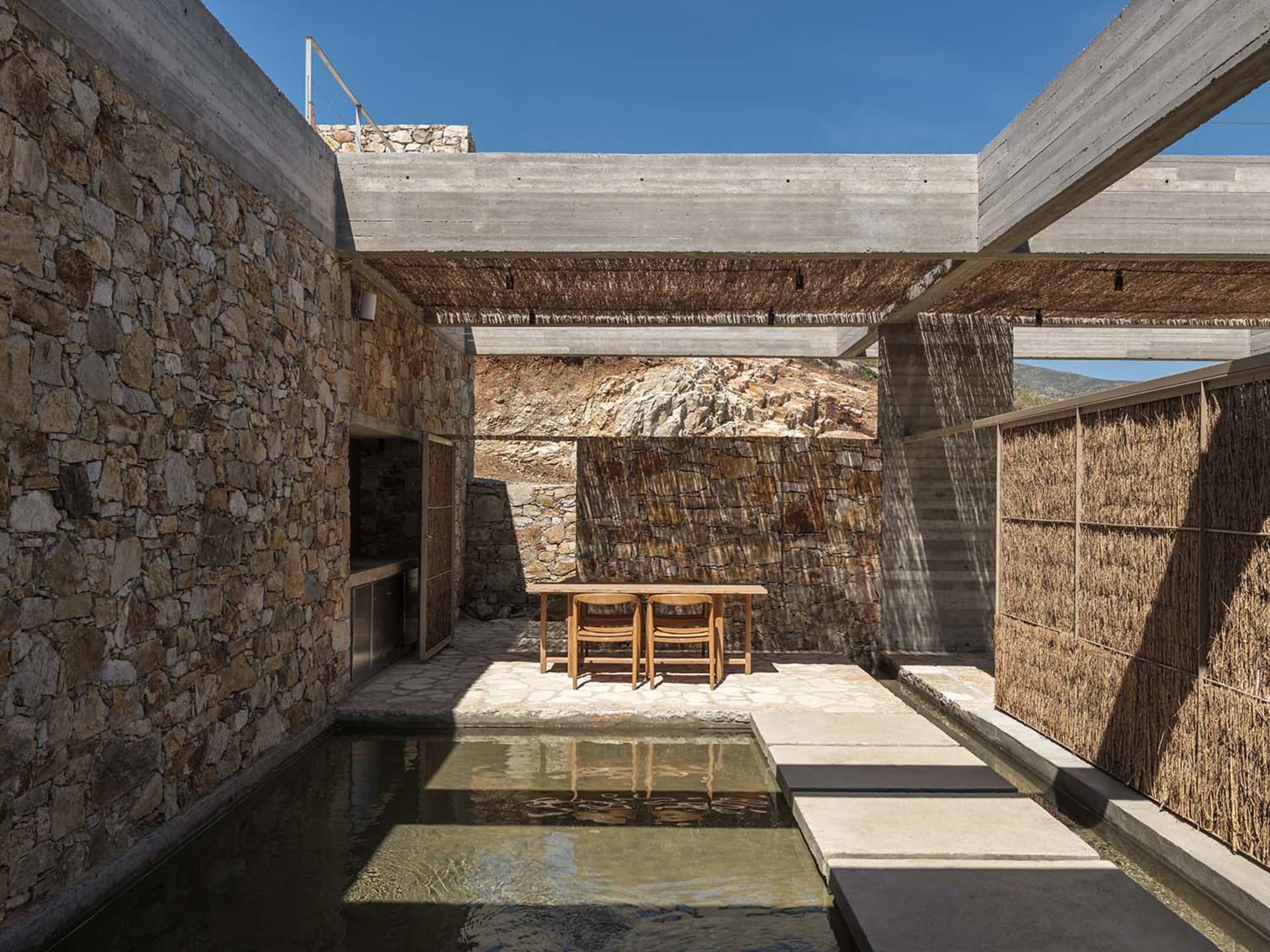Sunlit outdoor courtyard featuring irregular stone walls, concrete structural beams, a shallow reflecting pool, wooden dining table with two chairs, and woven reed screens under a clear blue sky.