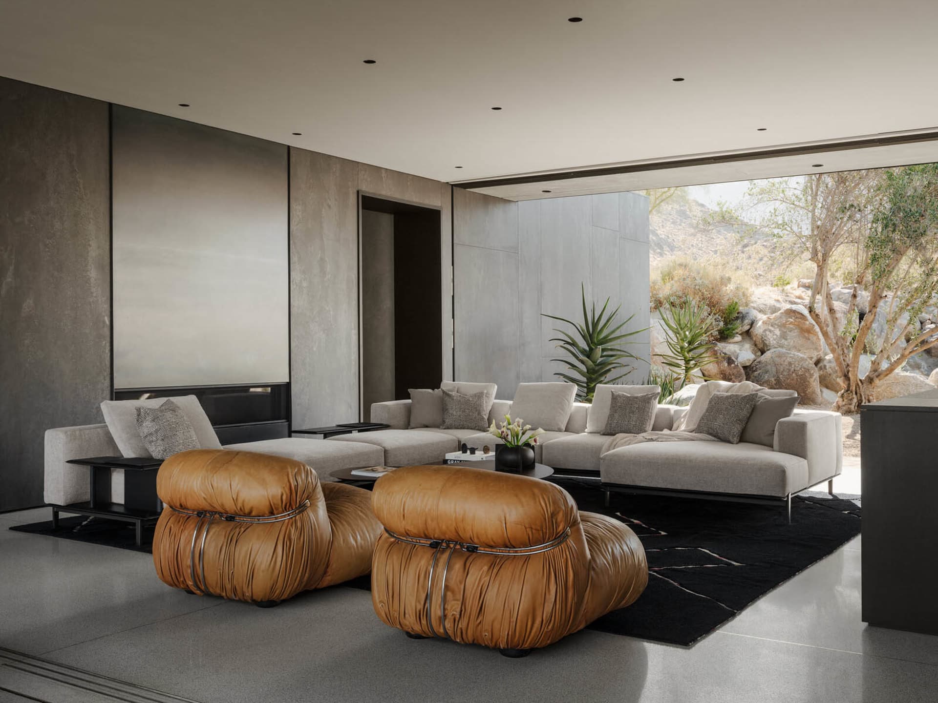Modern minimalist living room featuring two large brown leather poufs, a light grey sectional sofa, concrete walls, and large opening to desert landscape with rocks and trees.