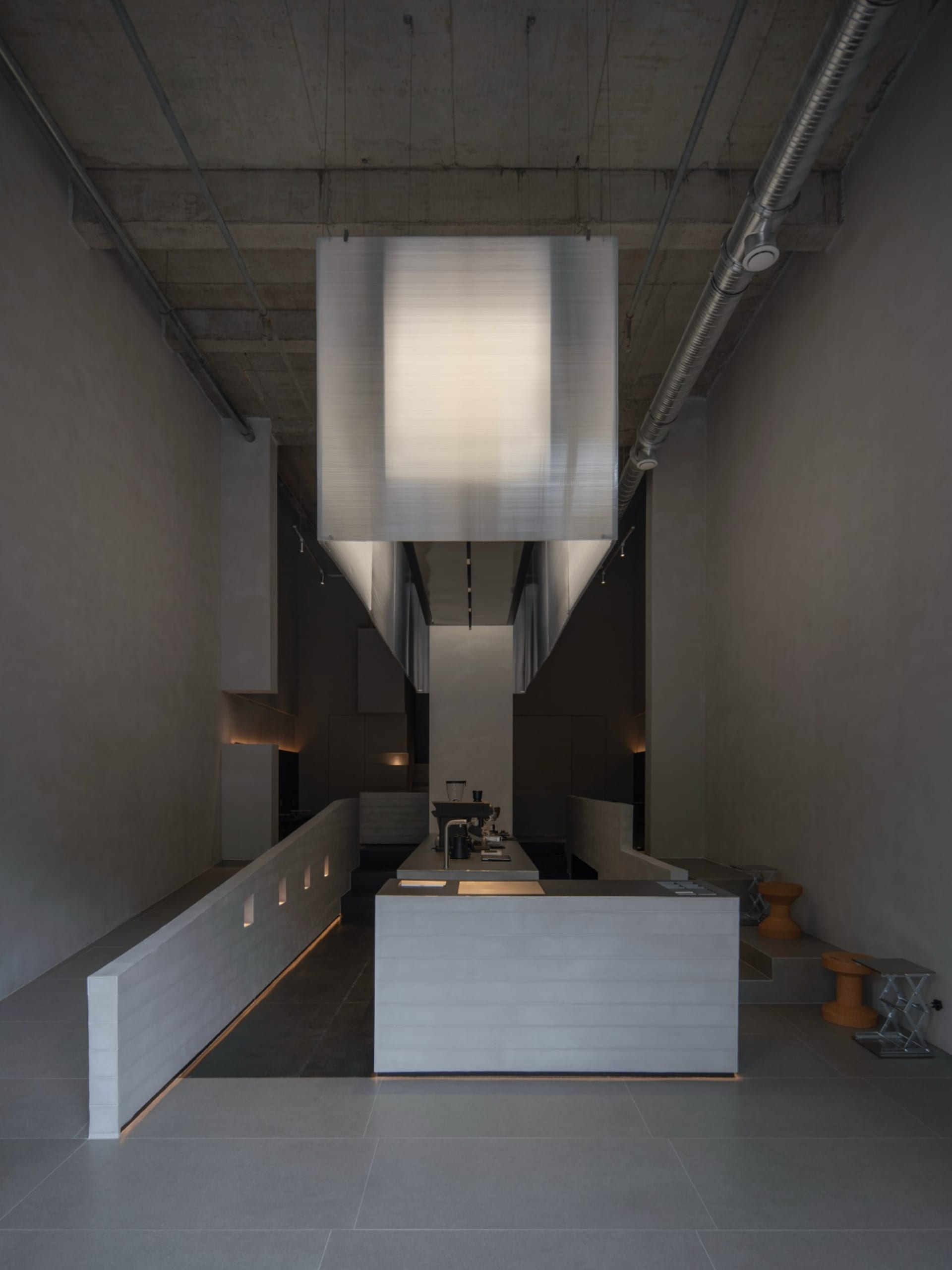 Modern brutalist kitchen interior featuring exposed concrete walls, floor, and ceiling, with a large rectangular fabric pendant light hanging over a concrete island, exposed metal ducts, and integrated LED lighting.