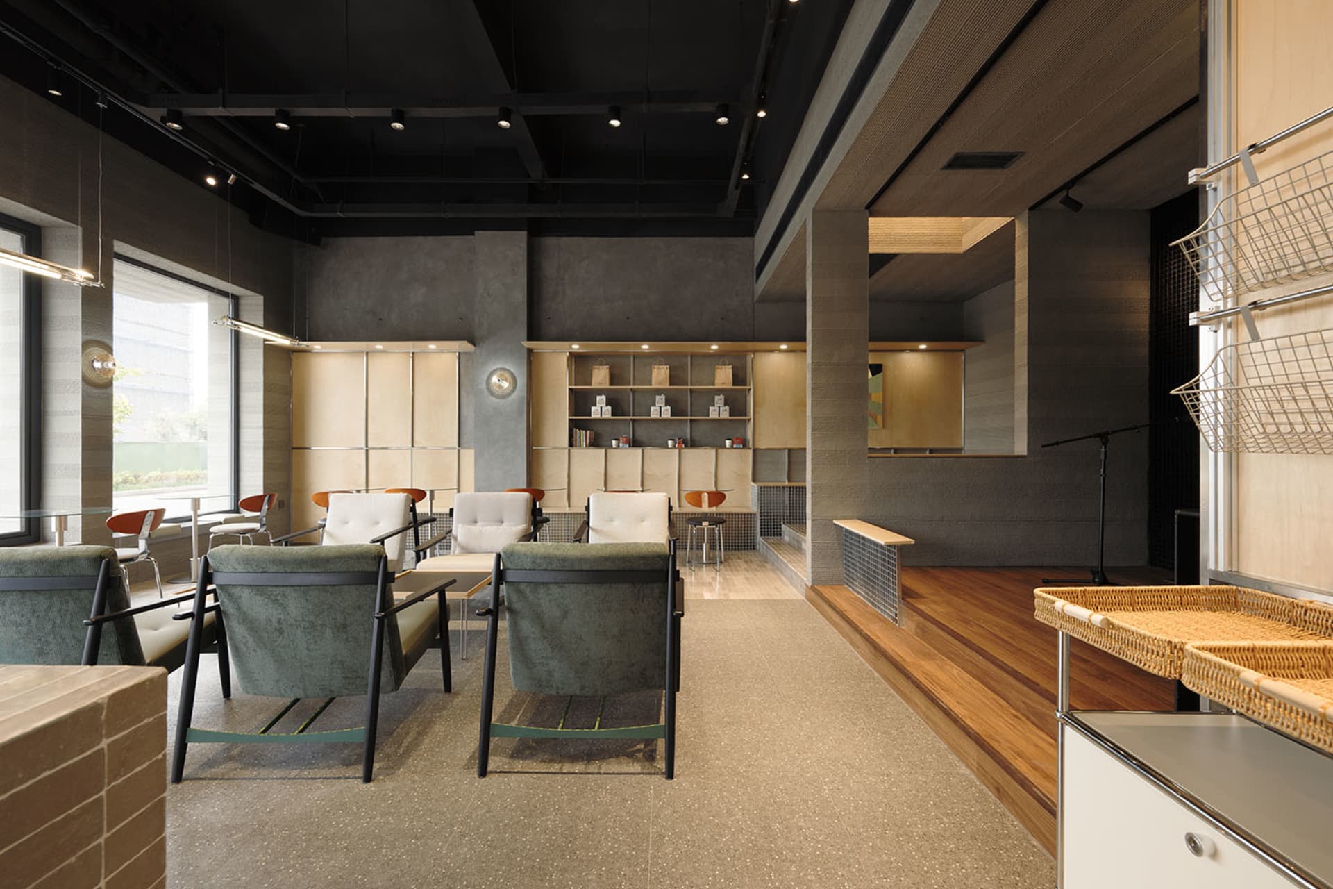 Modern retail lounge interior featuring green upholstered armchairs, concrete walls, warm wood flooring, and built-in light-wood display shelving.