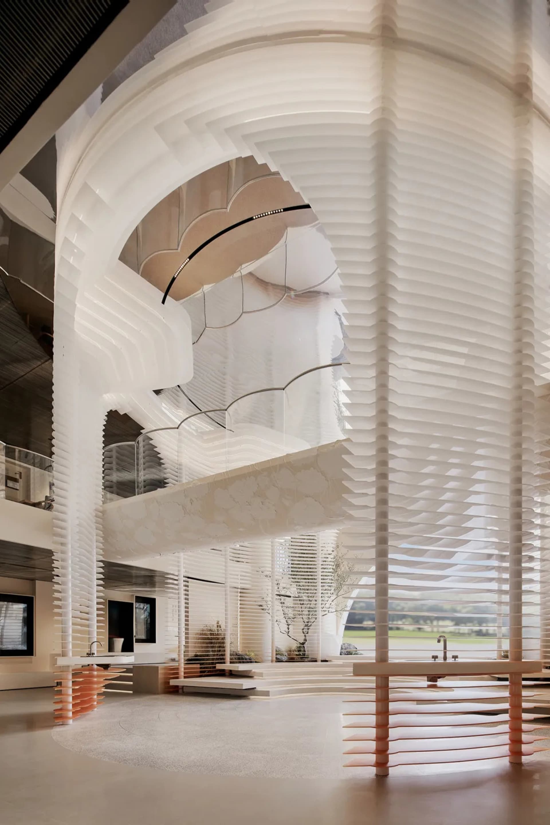 View of a modern multi-level atrium featuring white sculptural ribbed columns, layered cloud-like ceiling installations, and a minimalist open-plan space with natural light.