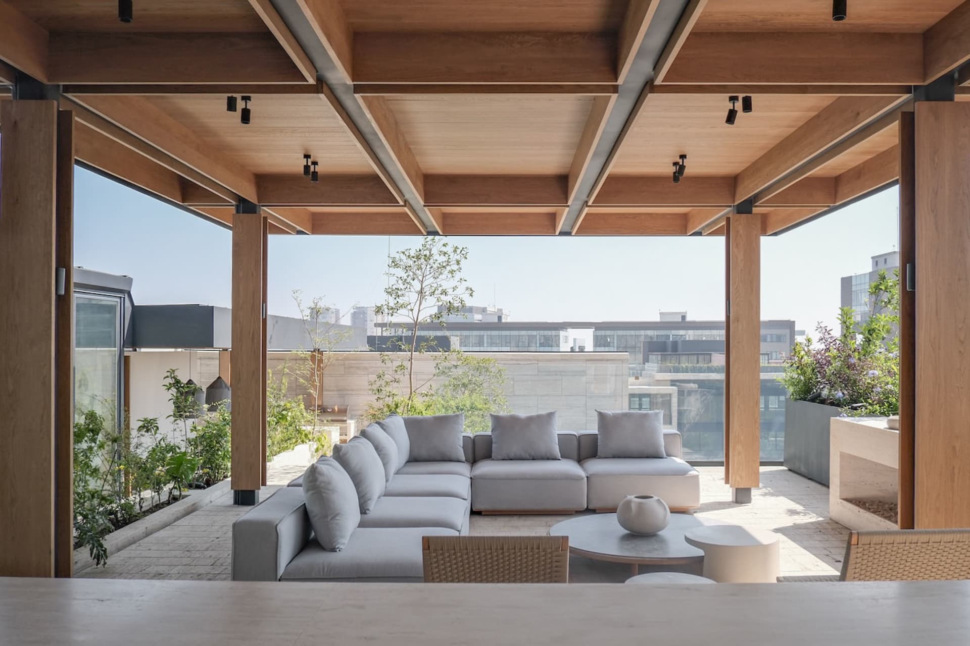 Modern rooftop terrace featuring a large grey L-shaped outdoor sofa, round concrete coffee tables, teak wood pergola with recessed lighting, and lush greenery overlooking city buildings.