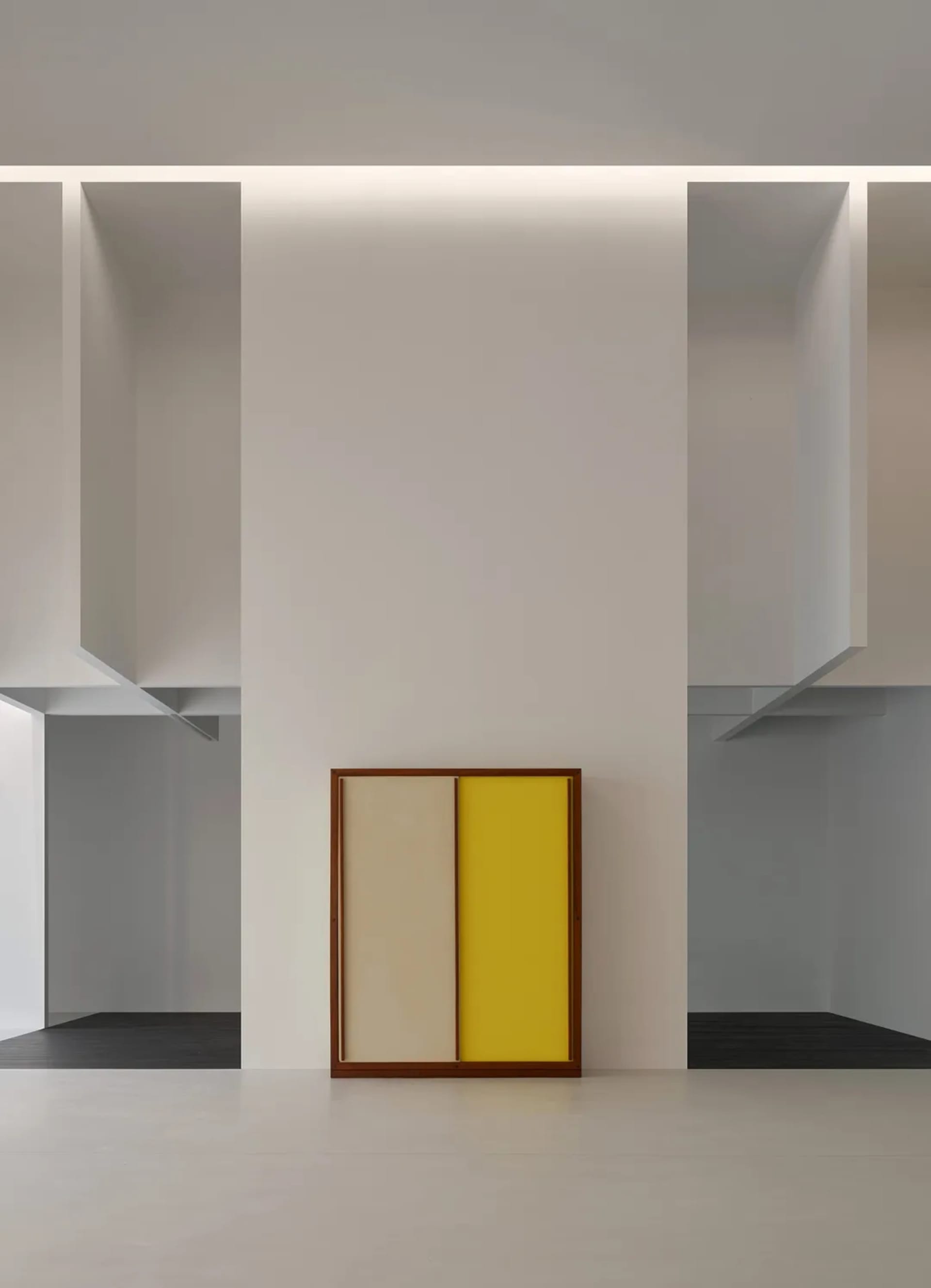 A framed two-tone panel with beige and bright yellow sections leaning against a white wall in a minimalist gallery space with dramatic architectural cutouts and hidden lighting.