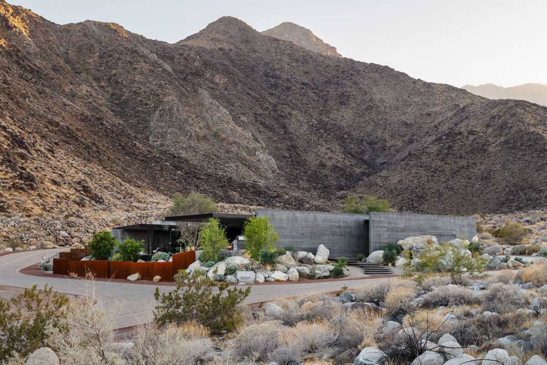 Modern concrete house with large glass windows nestled among desert rocks and native plants, with a massive arid mountain range in the background at golden hour.