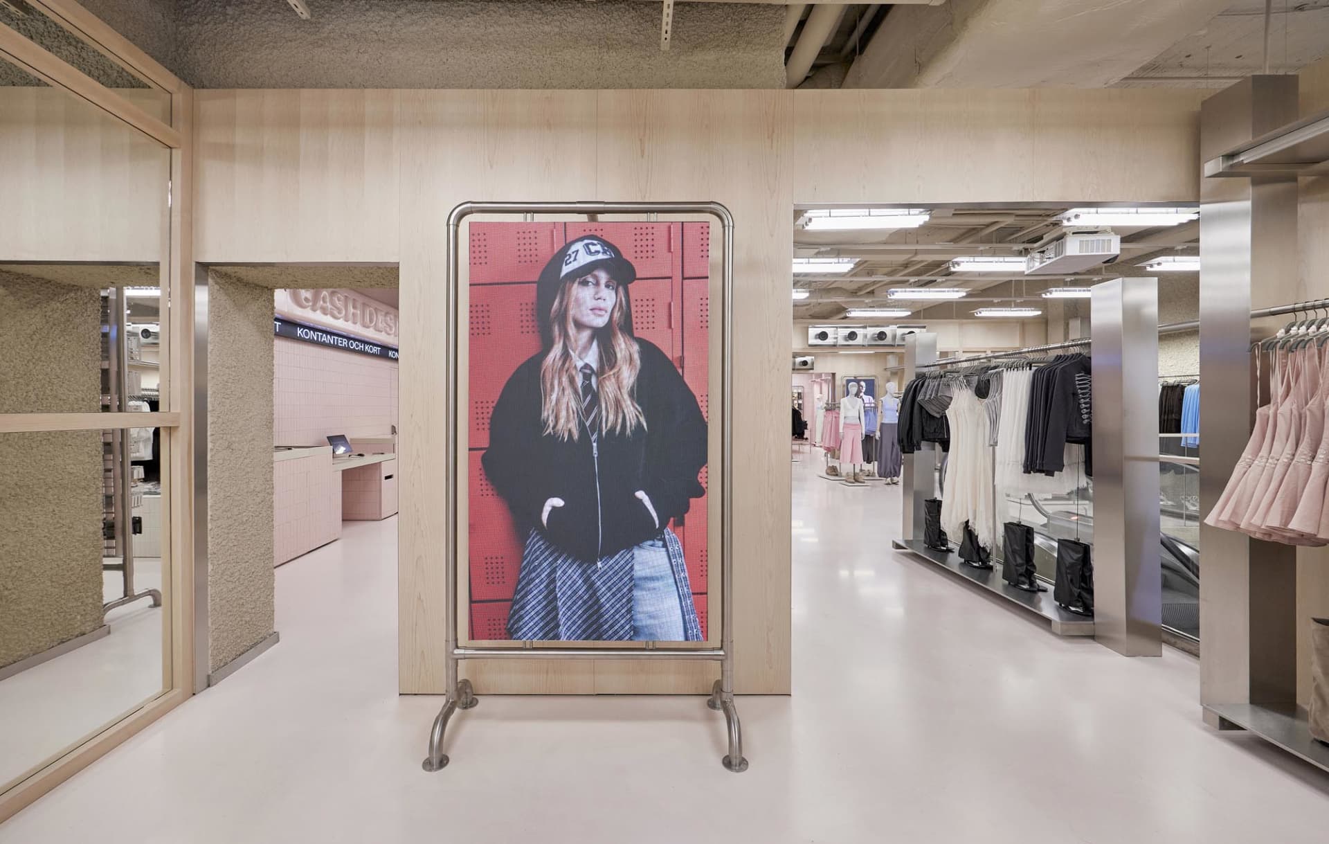 Modern fashion boutique interior featuring light wood paneling, a large digital portrait on a metal stand, stainless steel accents, and clothing racks in the background.