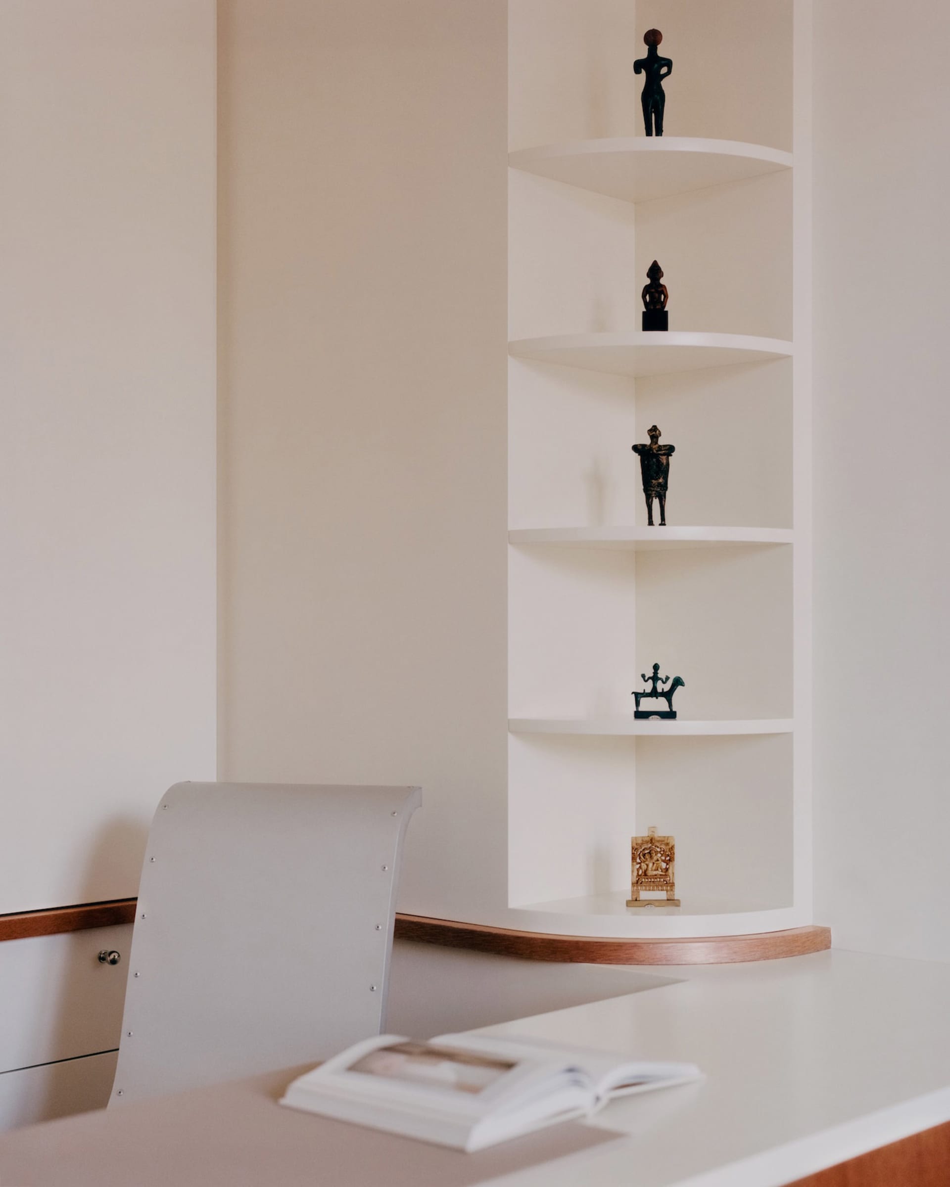Modern minimalist white corner built-in shelves displaying four African tribal sculptures above a white desk with an open book and light grey chair.