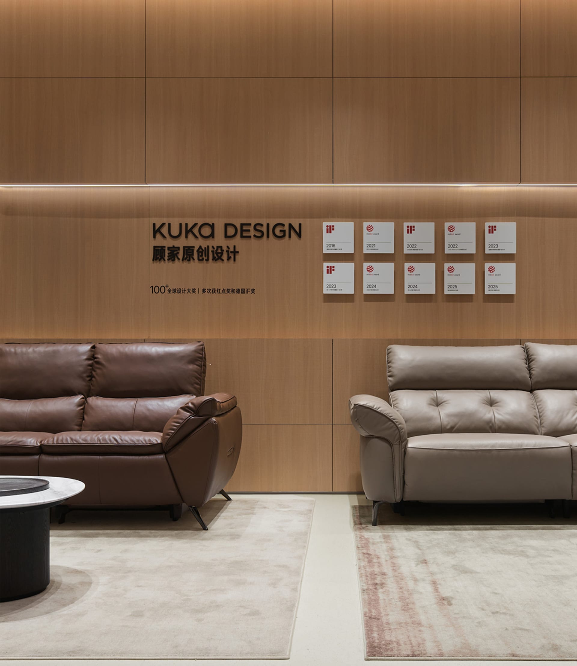 Modern showroom interior with a brown leather sofa on the left and a beige leather sofa on the right, set against a backdrop of light wood paneling with KUKA DESIGN branding and multiple iF and Red Dot design awards displayed.