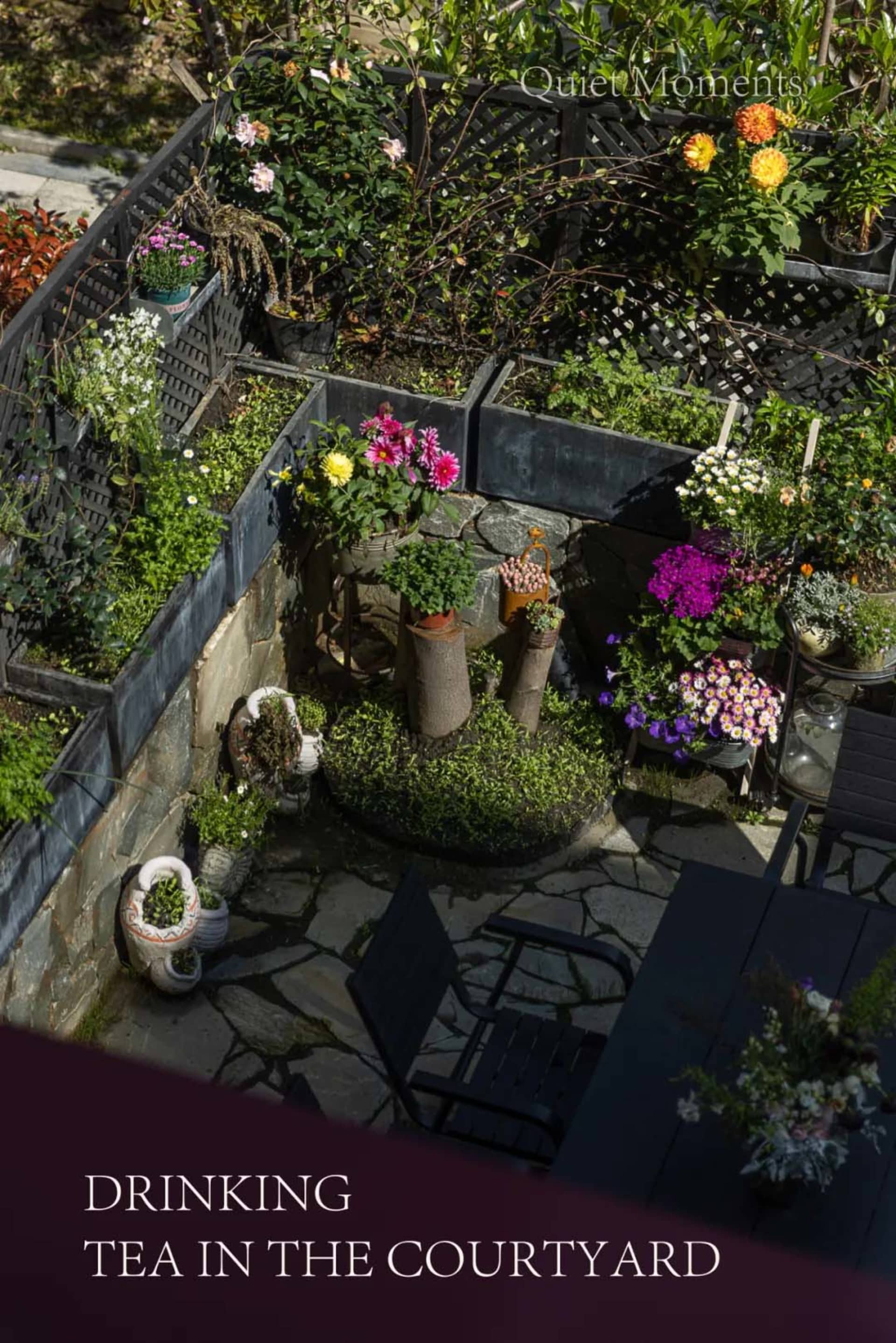 Overhead view of a charming stone-paved courtyard garden featuring abundant colorful potted flowers, wooden lattice planters, natural stone walls, and dark wooden outdoor furniture under bright daylight.