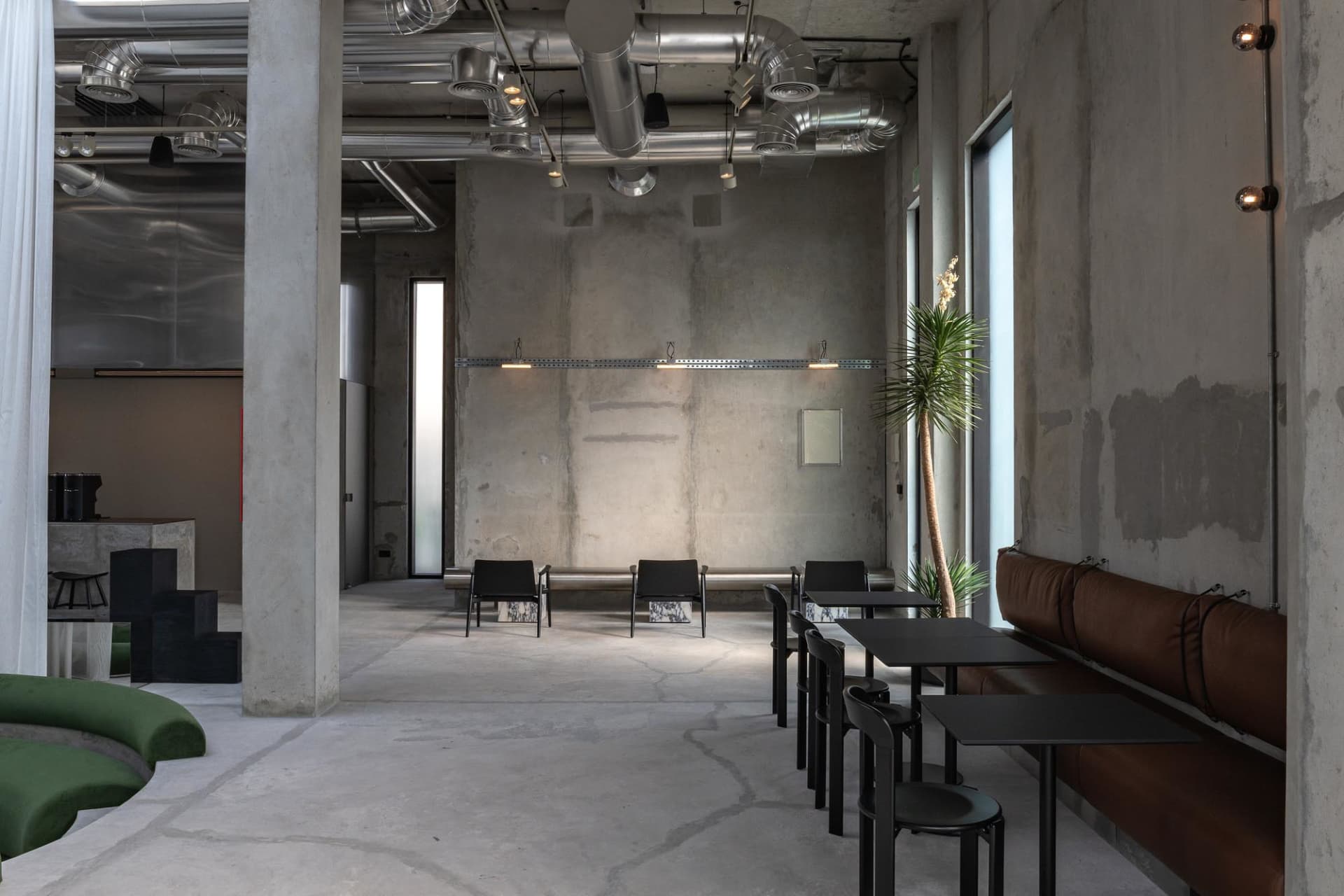 Spacious industrial-style cafe interior with exposed concrete walls, silver ventilation ducts, brown leather banquette seating, black dining tables, and a curved green modular sofa in the foreground.