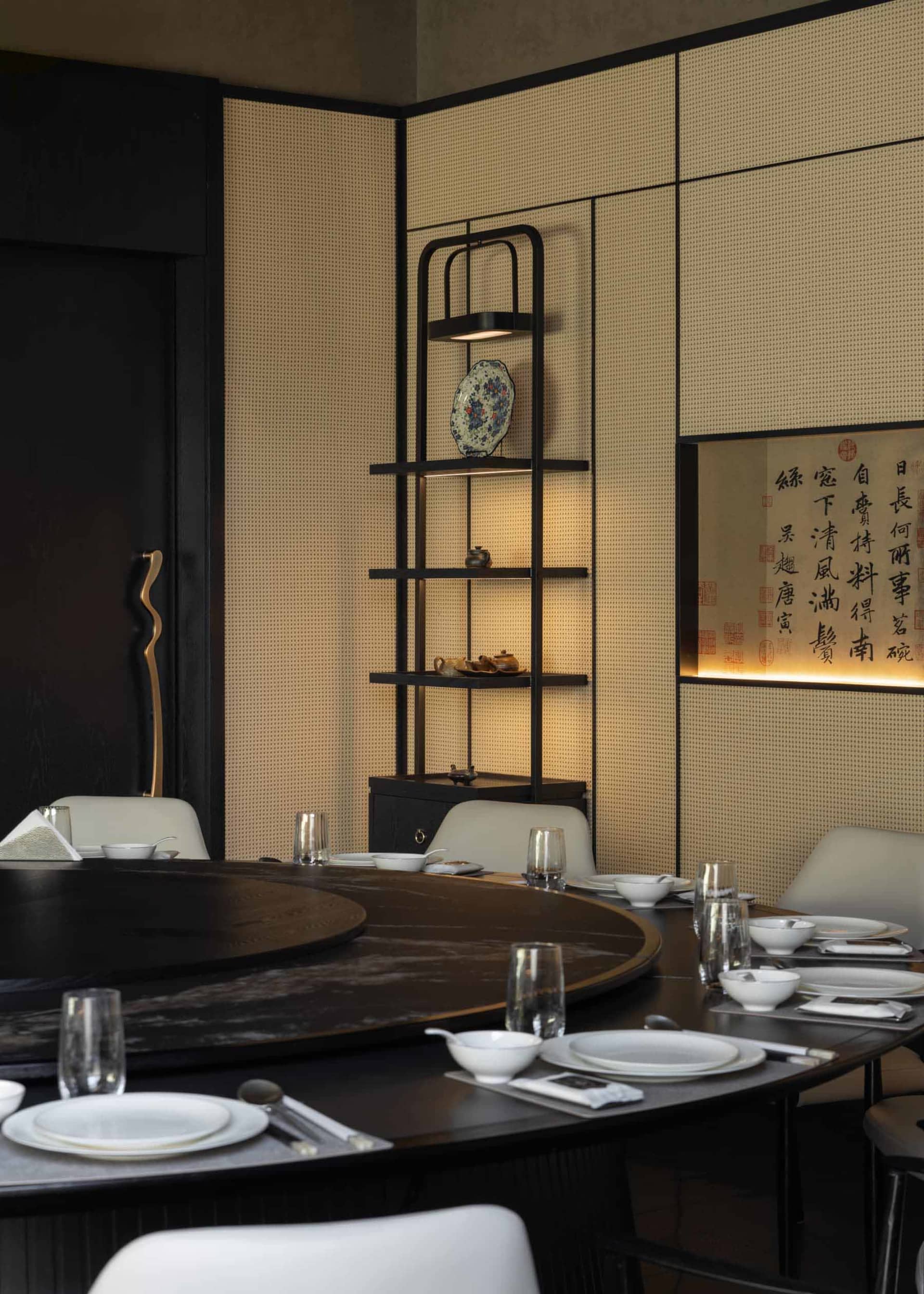 Modern Chinese-style dining room featuring a large round walnut table, white upholstered chairs, illuminated lattice wall panels, and a black metal display shelf with decorative ceramics and calligraphy art.