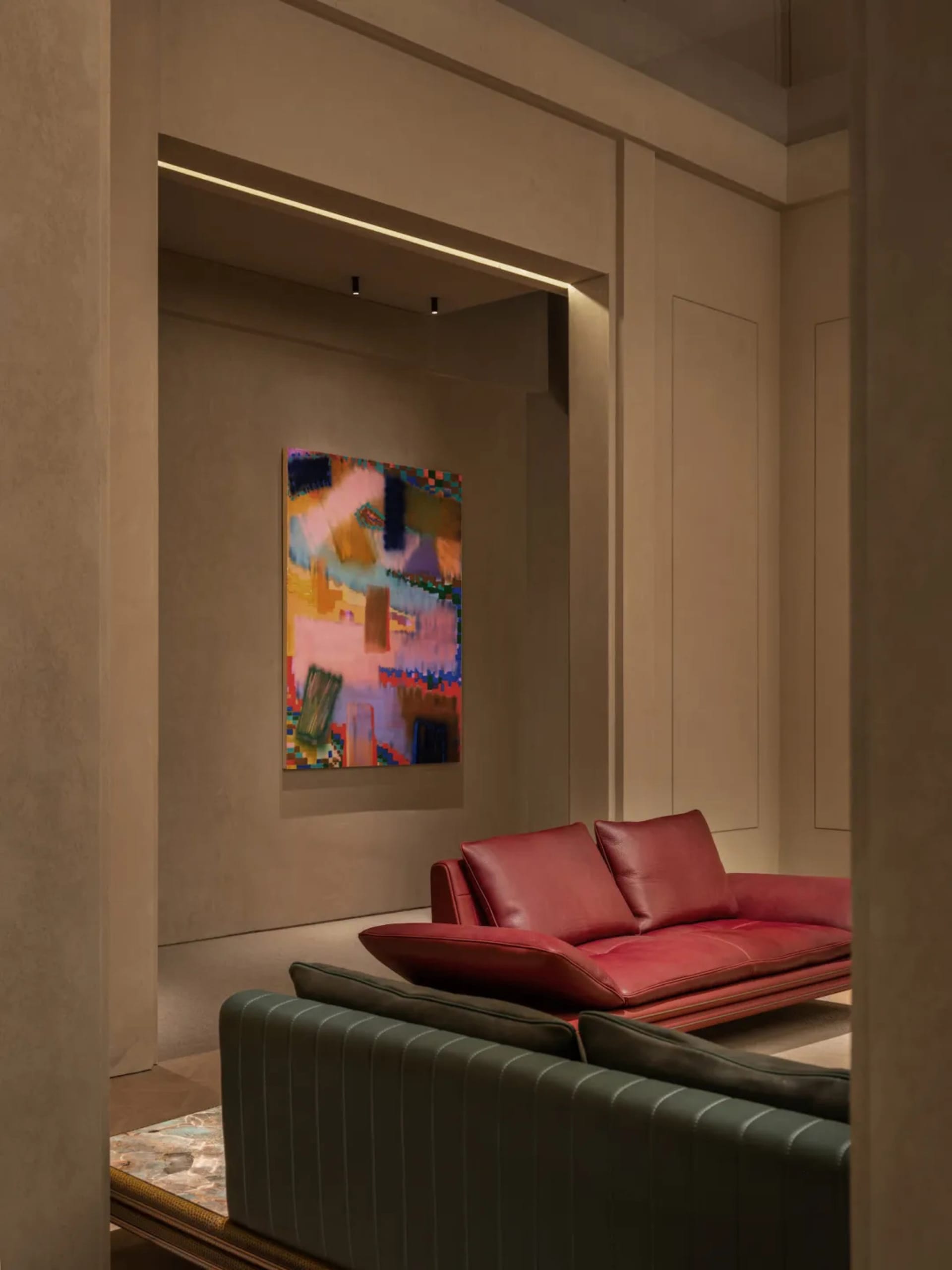 Modern living room corner featuring a red leather sofa, dark green striped armchair, and a large colorful abstract painting on a textured beige wall.