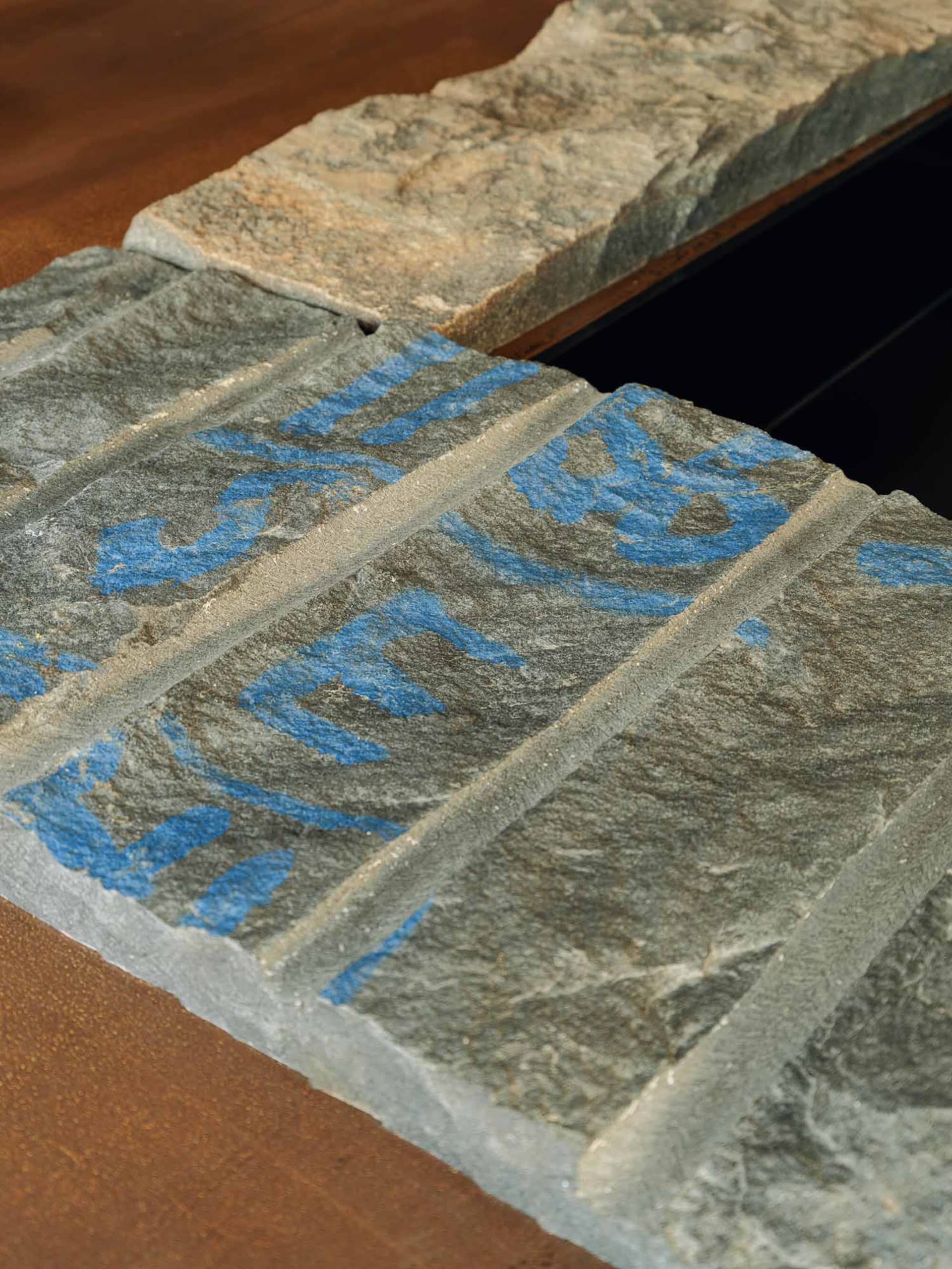 Close-up photograph of a thick natural slate tabletop with prominent blue mineral streaks and rough textured surface, resting on a solid walnut wooden base.