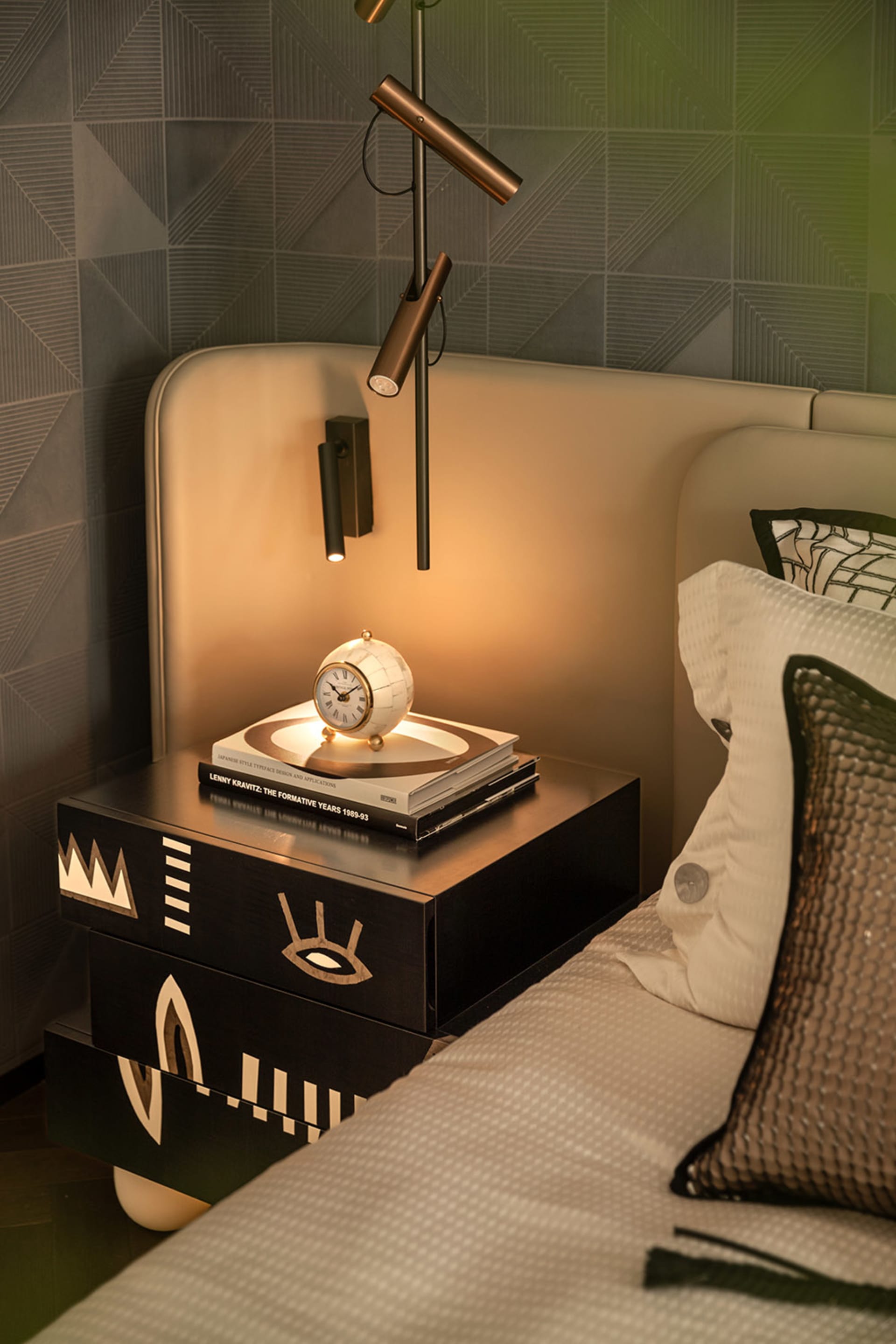 Close-up of a modern bedroom corner featuring a black lacquered bedside table with gold abstract inlays, a spherical clock, stacked design books, and contemporary wall sconces against a geometric patterned wall.