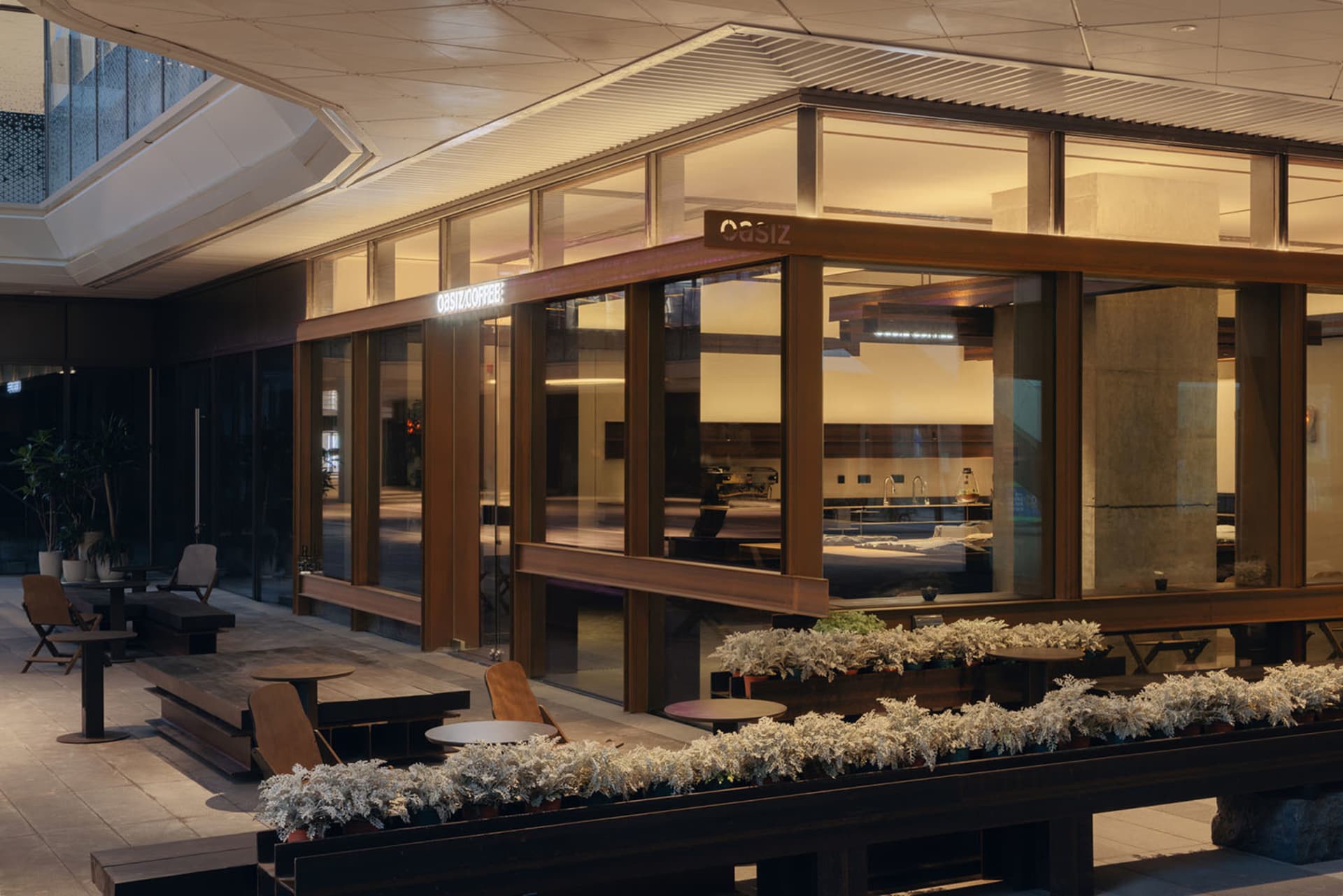 Modern outdoor café terrace at night featuring a glass and wood pavilion, wooden seating, and white-flowered planters under warm lighting.