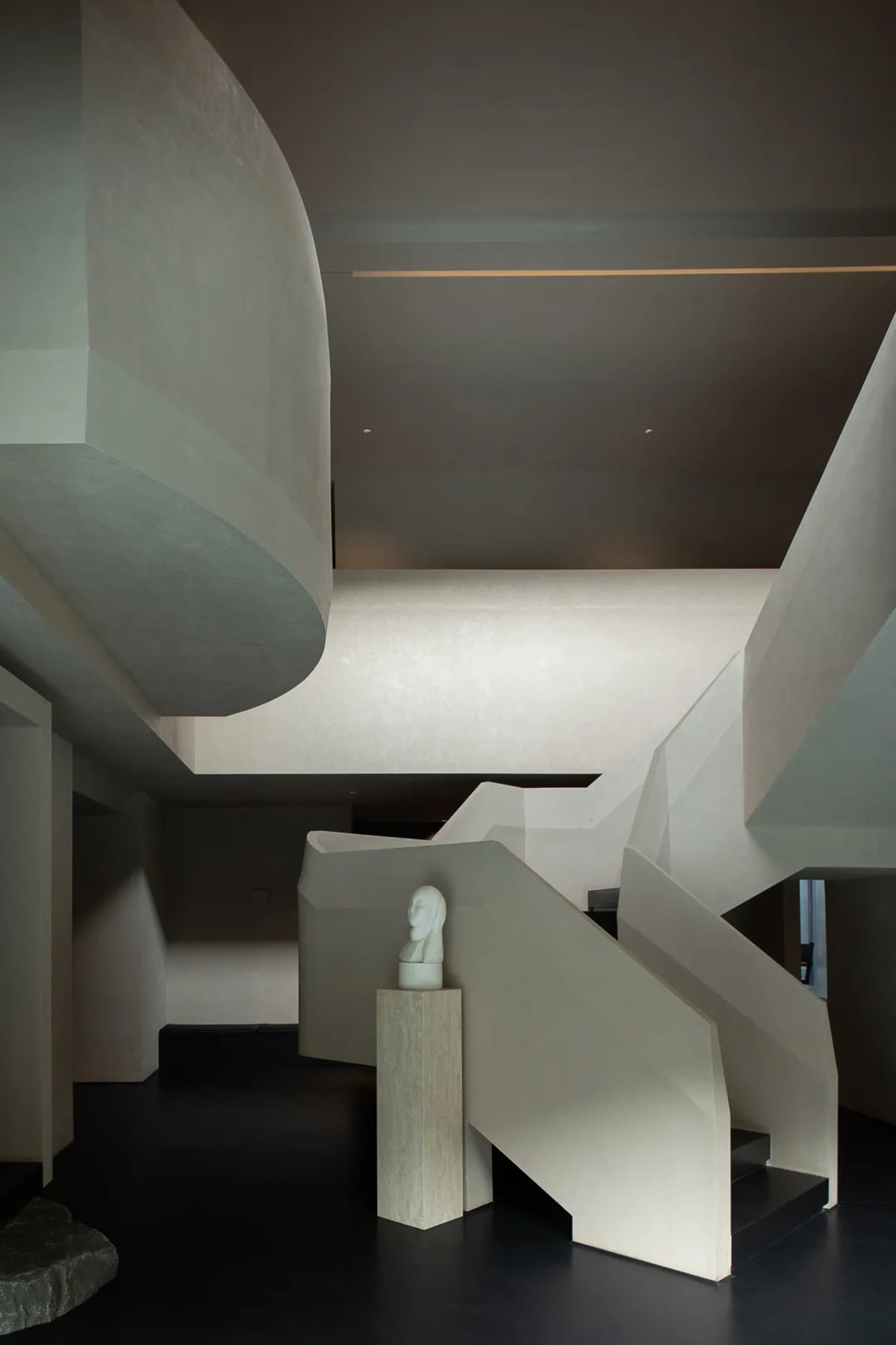 Modern sculptural concrete staircase with integrated lighting, featuring a white marble bust on a stone pedestal against a dark floor in a minimalist interior.