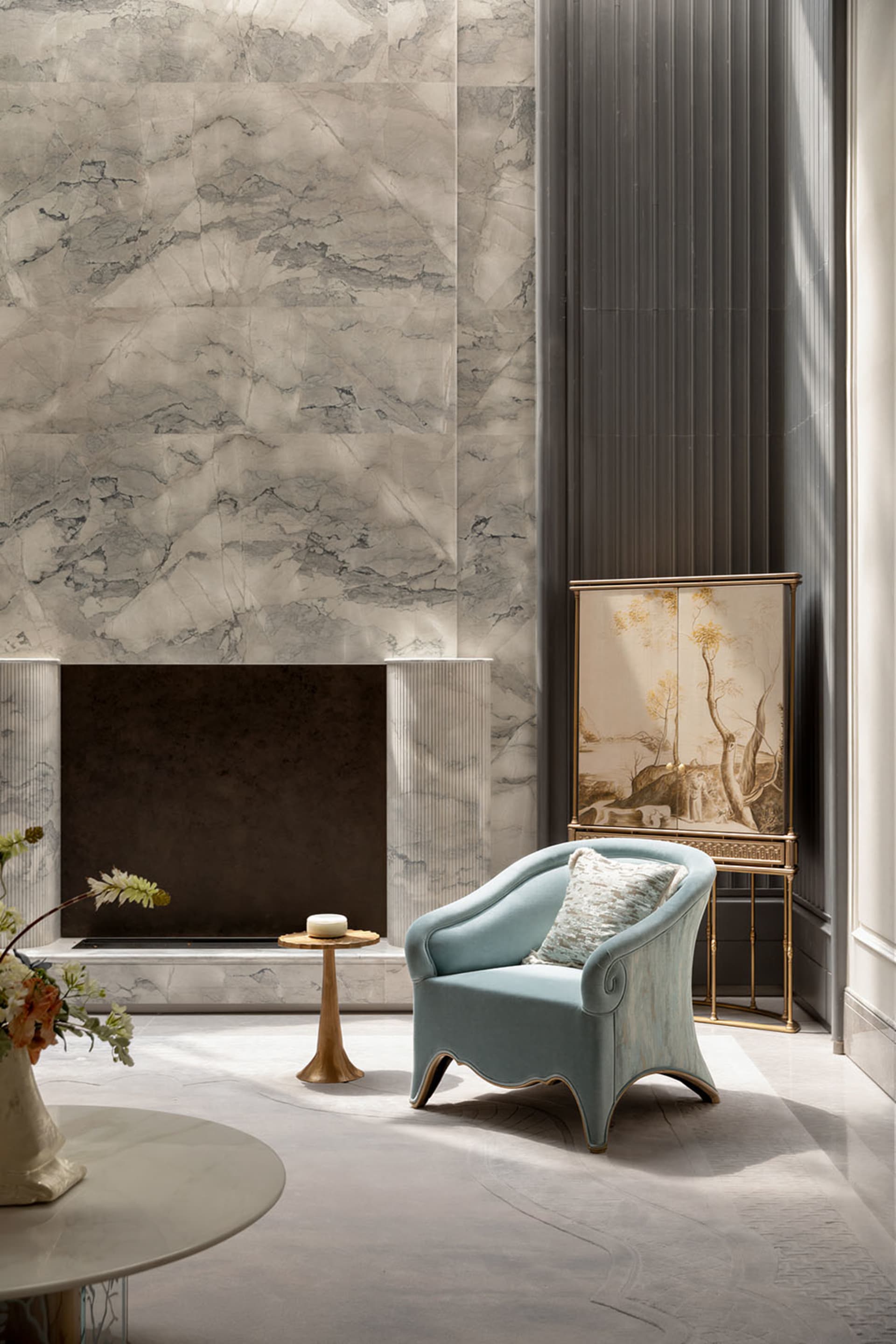Elegant contemporary living room featuring a large marble fireplace, a curved light-blue velvet armchair with patterned cushion, a golden side table, and a traditional Chinese painted folding screen against a dark fluted wall panel.
