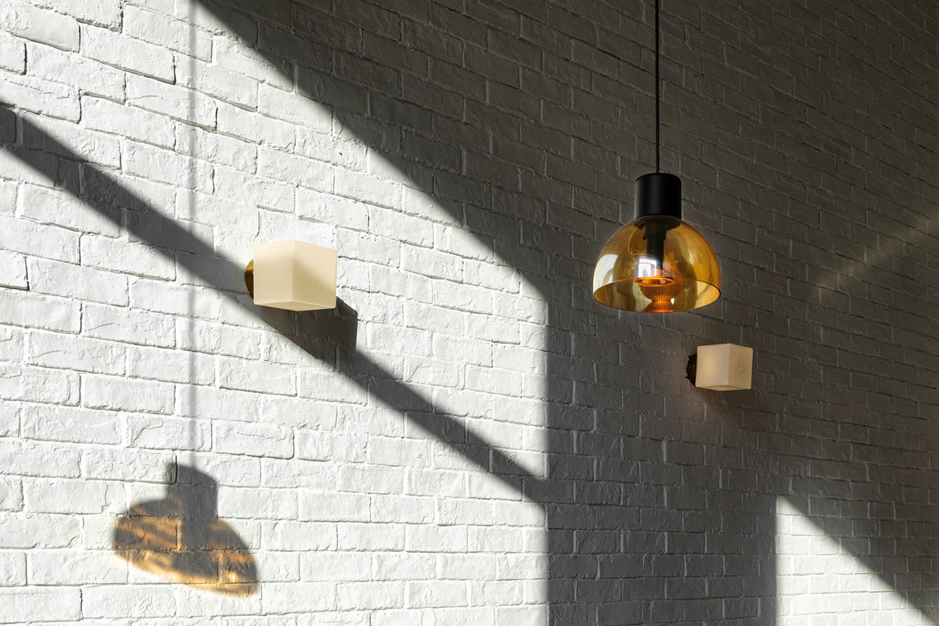 White painted brick wall illuminated by dramatic diagonal sunlight, featuring a large amber glass pendant lamp and two small cube-shaped wall sconces casting strong geometric shadows.