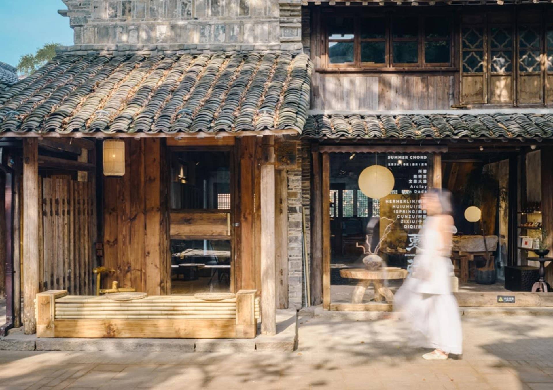 Traditional Chinese wooden shopfront with grey tiled roof, large timber pillars, and a blurred figure in white walking past on a sunny day.