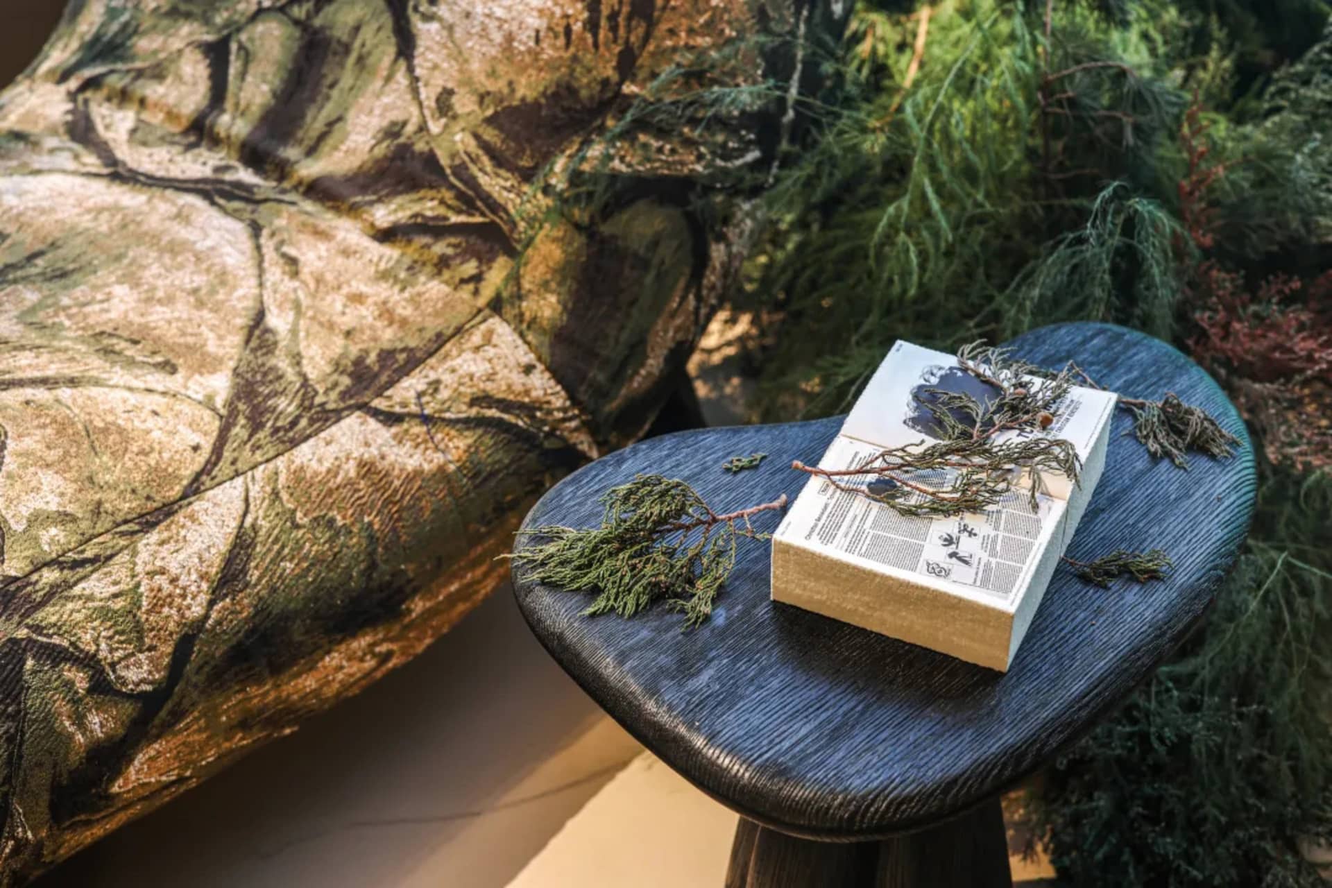 Close-up of a dark charred wood side table holding an open book and cypress branches, next to a large sofa upholstered in a realistic forest moss and tree bark pattern.