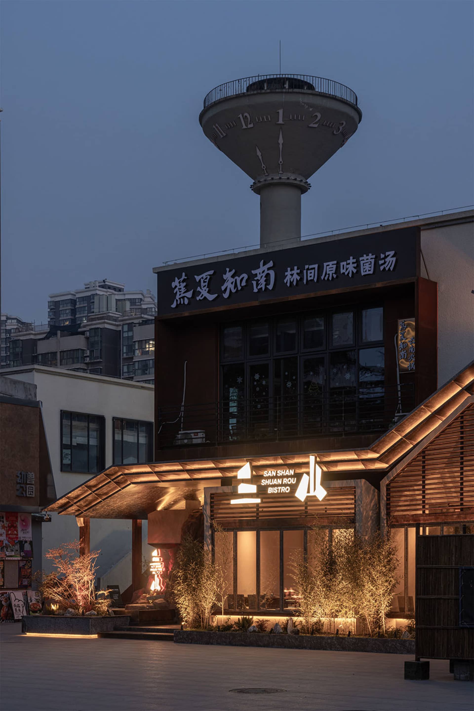 Night view of San Shan Shuan Rou Bistro with illuminated corten steel facade, wooden slats, and lush landscaping under a large concrete water tower.