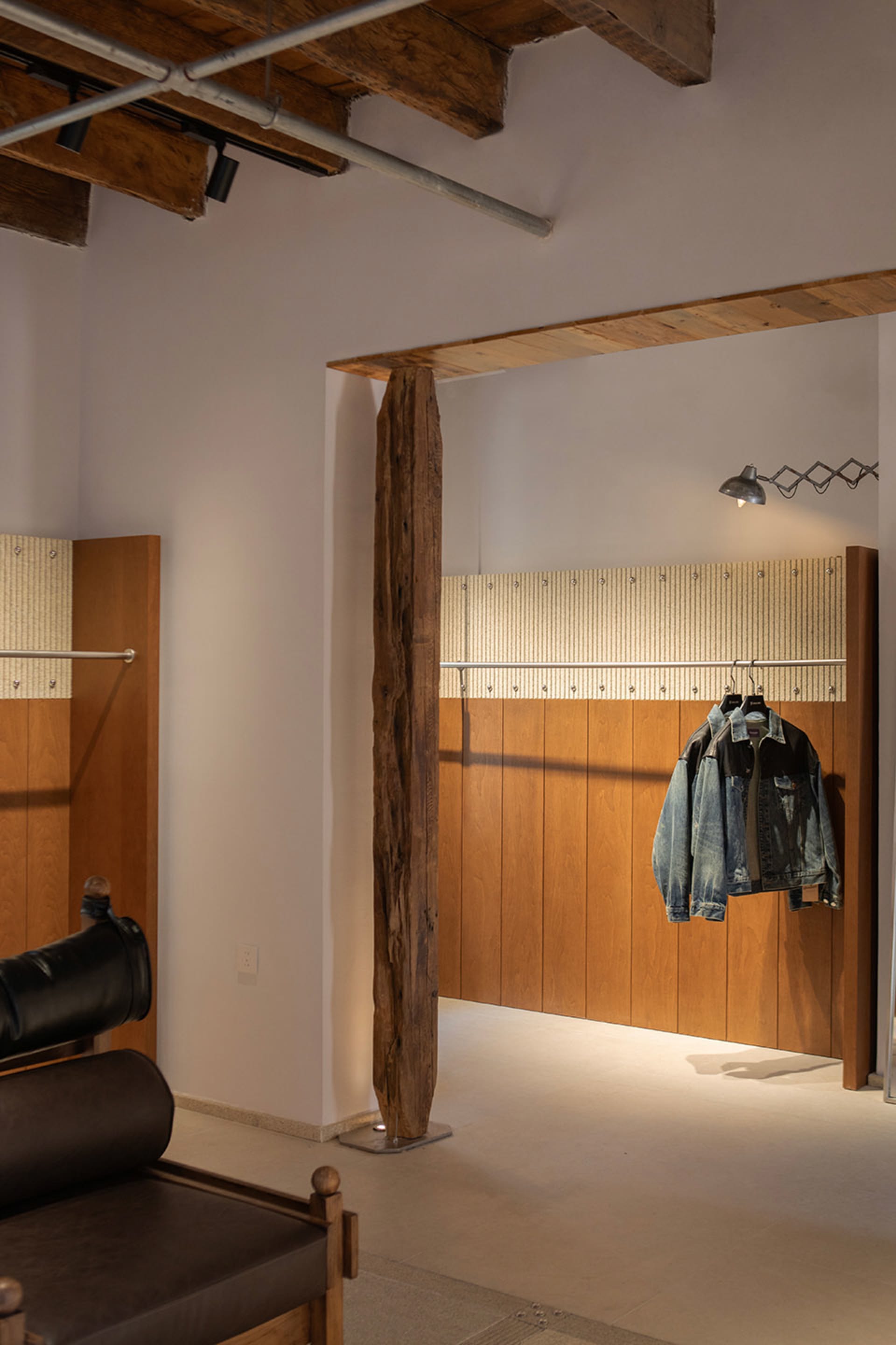 Modern industrial entryway featuring a tall reclaimed wood beam, warm vertical wood paneling, exposed ceiling pipes, and two denim jackets hanging on a metal rod under a wall lamp.