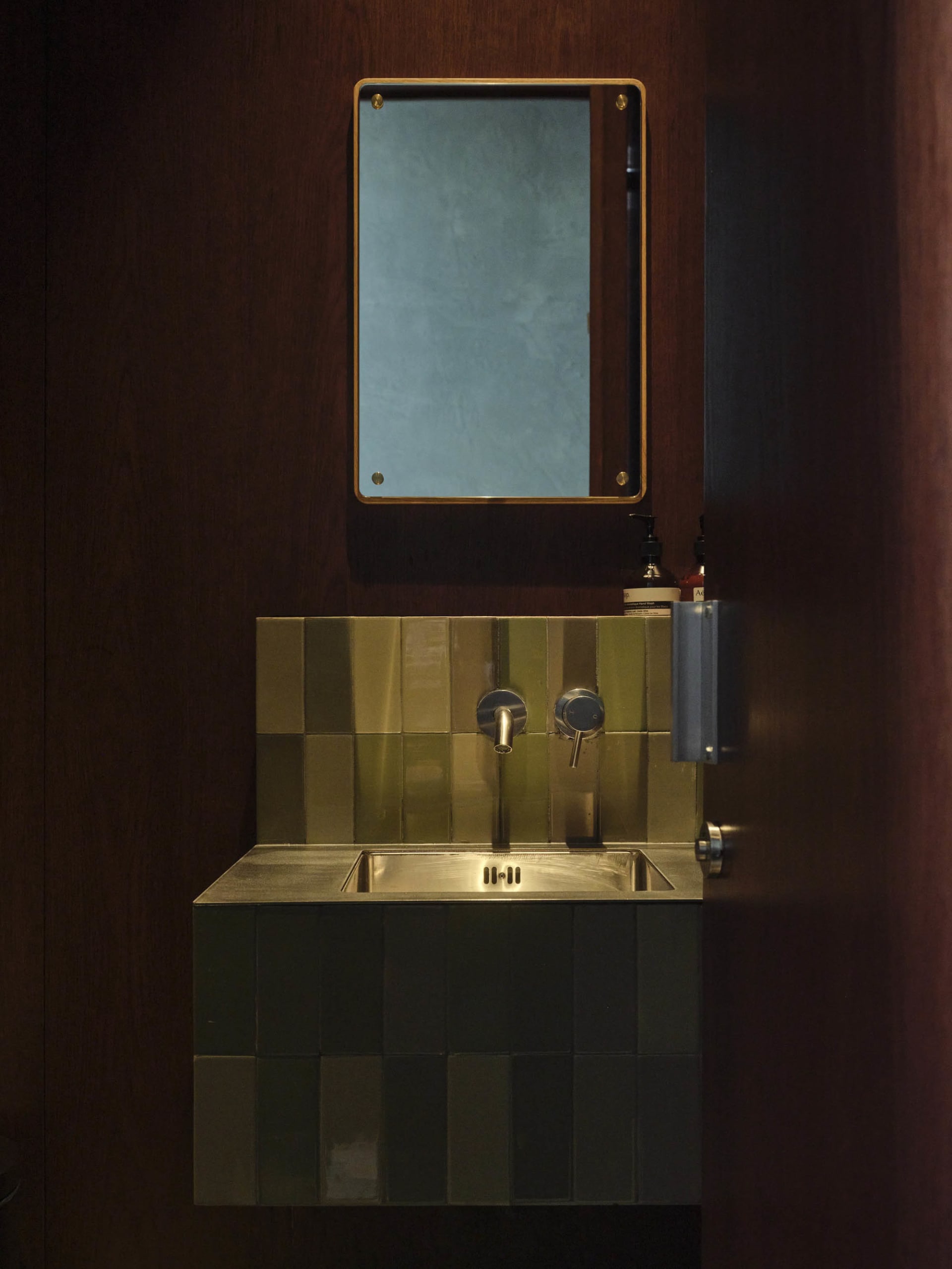 Wall-mounted stainless steel utility sink with green and olive checkerboard tiles, brass fixtures, and a rectangular mirror against dark wood walls.