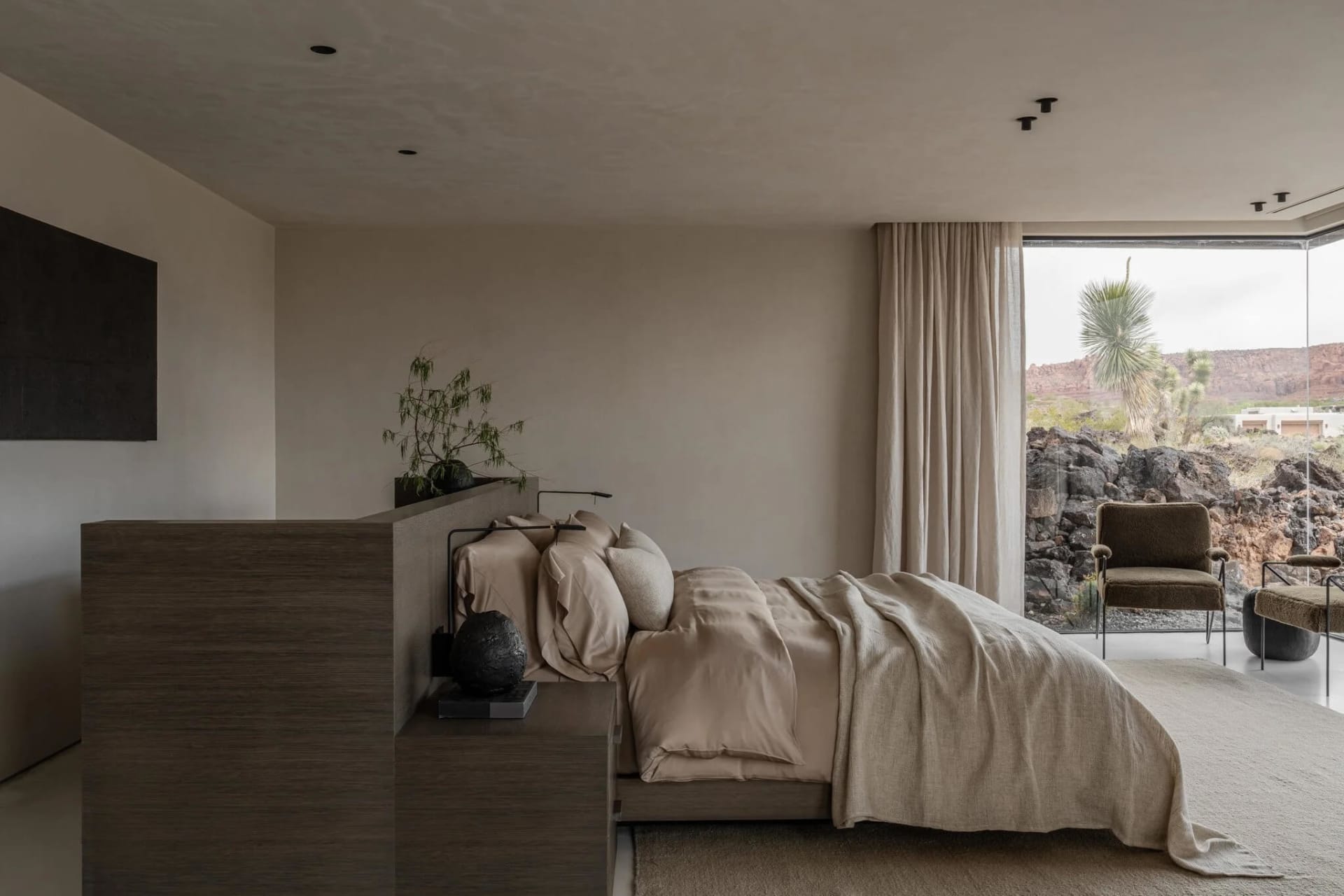 Modern minimalist bedroom featuring a low wooden platform bed with beige linen bedding, textured wood headboard, and large glass doors opening to a rocky desert landscape with yucca plants.