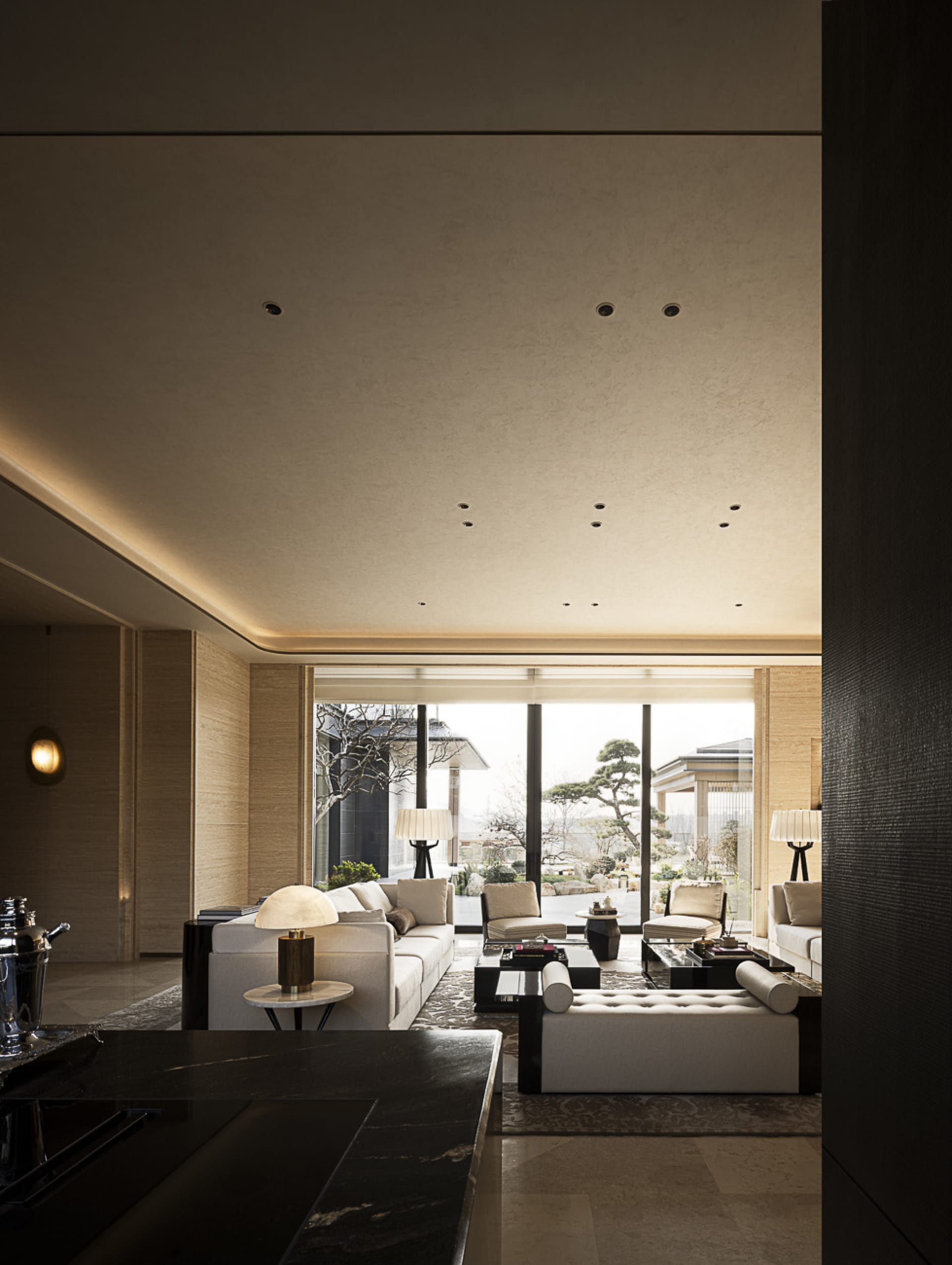Modern luxury living room featuring travertine walls, large white sectional sofa, dark marble coffee tables, and floor-to-ceiling windows overlooking a Japanese garden with bonsai trees.