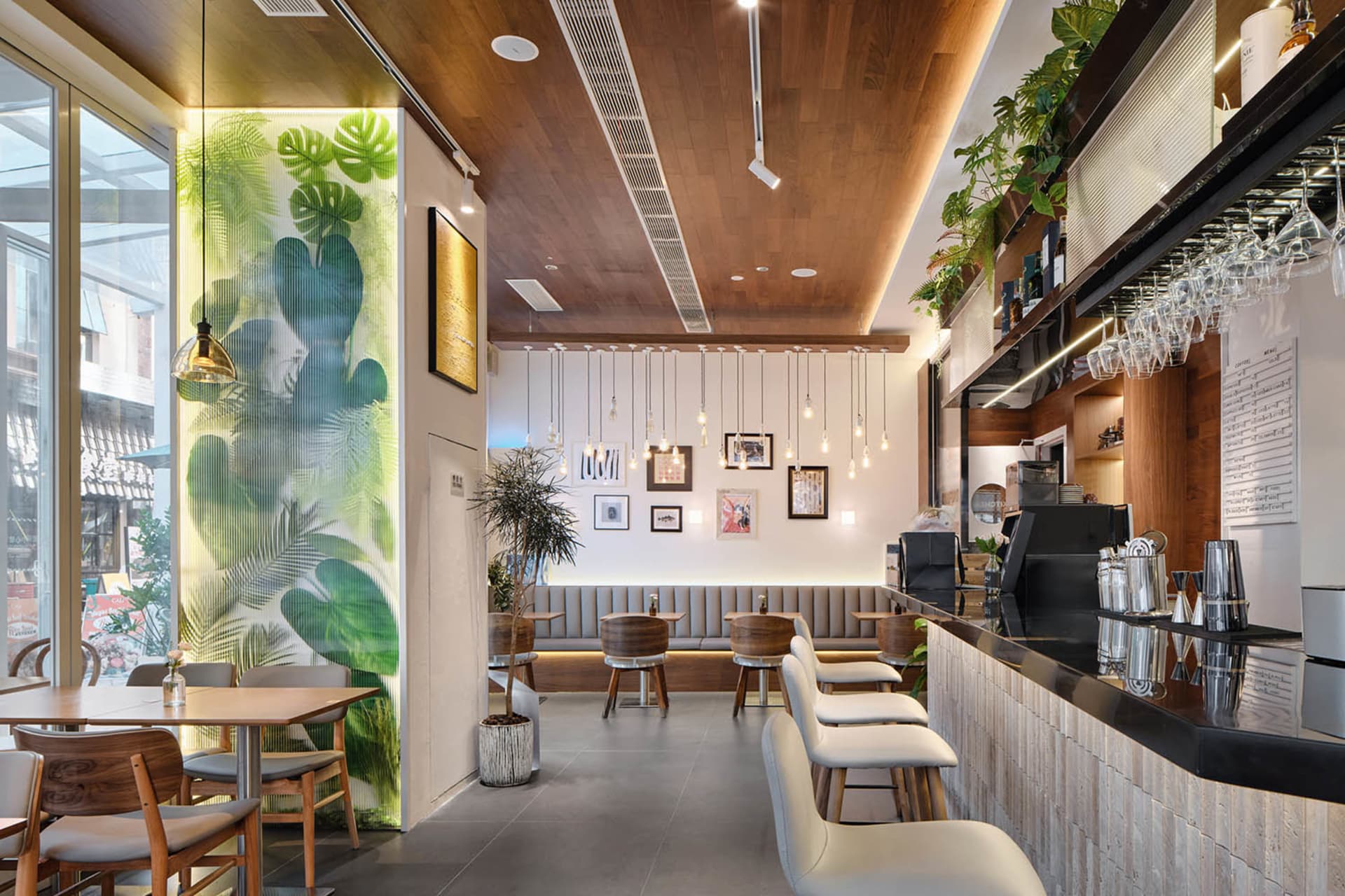 Modern cafe interior with a glowing green backlit monstera leaf feature wall, wooden ceiling and bar counter, white bar stools, and pendant lighting.