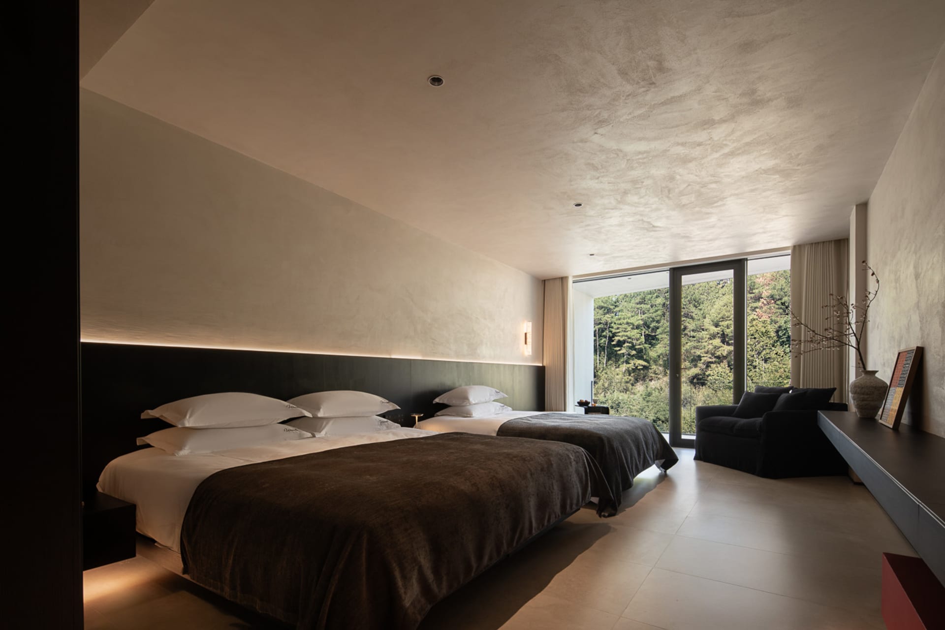 Modern minimalist twin hotel bedroom featuring two large beds with dark brown throws, a black upholstered headboard with LED backlighting, textured plaster walls, and large windows overlooking a dense forest.