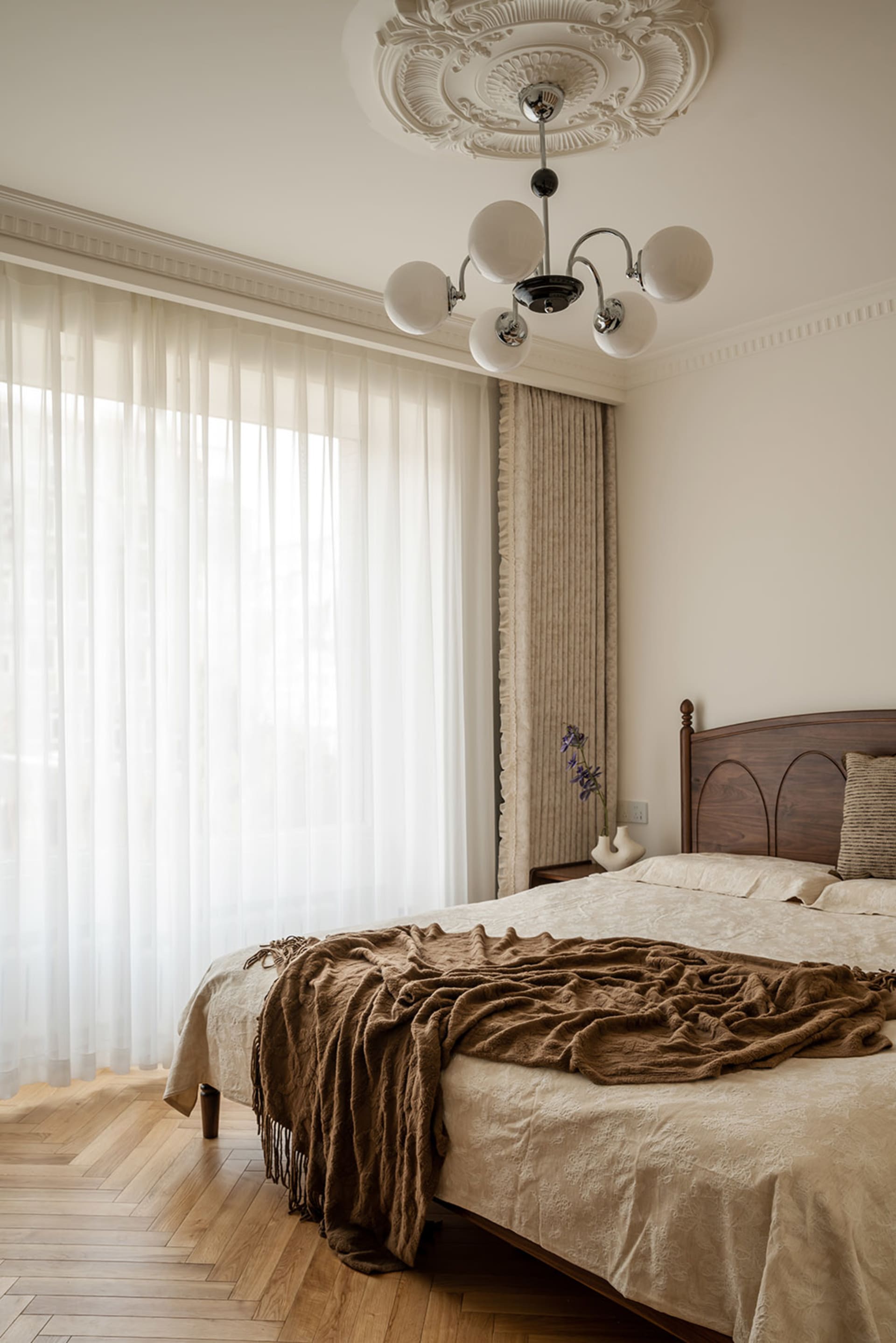 A tranquil bedroom featuring a dark walnut wooden bed, ornate white ceiling medallion with modern chandelier, sheer white curtains, textured beige drapes, and a bed layered with cream patterned linens and a rumpled brown throw blanket on herringbone wood flooring.