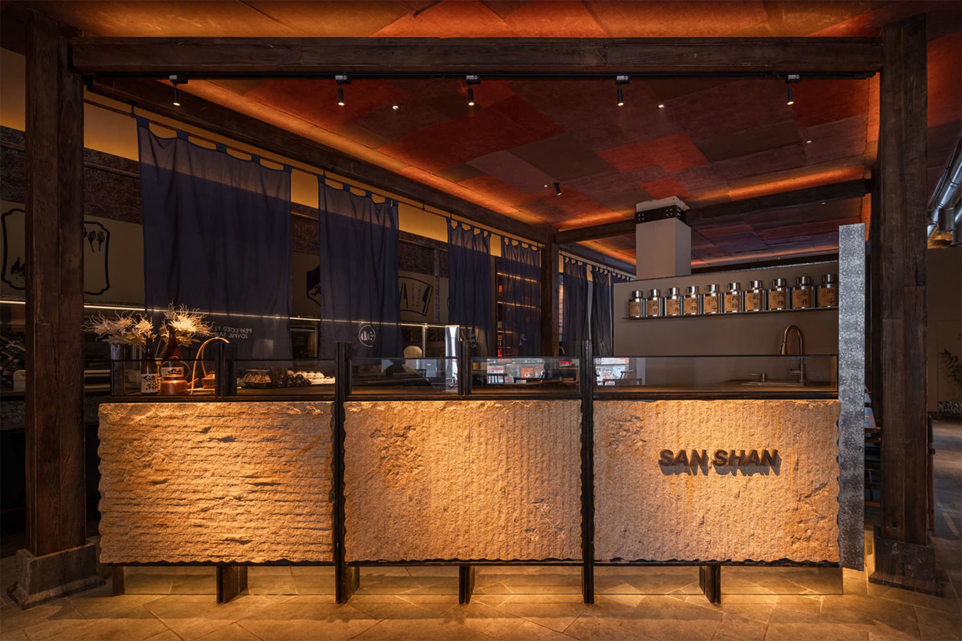 Modern Japanese-inspired cafe interior featuring a massive textured stone counter with backlit 'SAN SHAN' signage, dark timber structural beams, and warm ambient lighting.