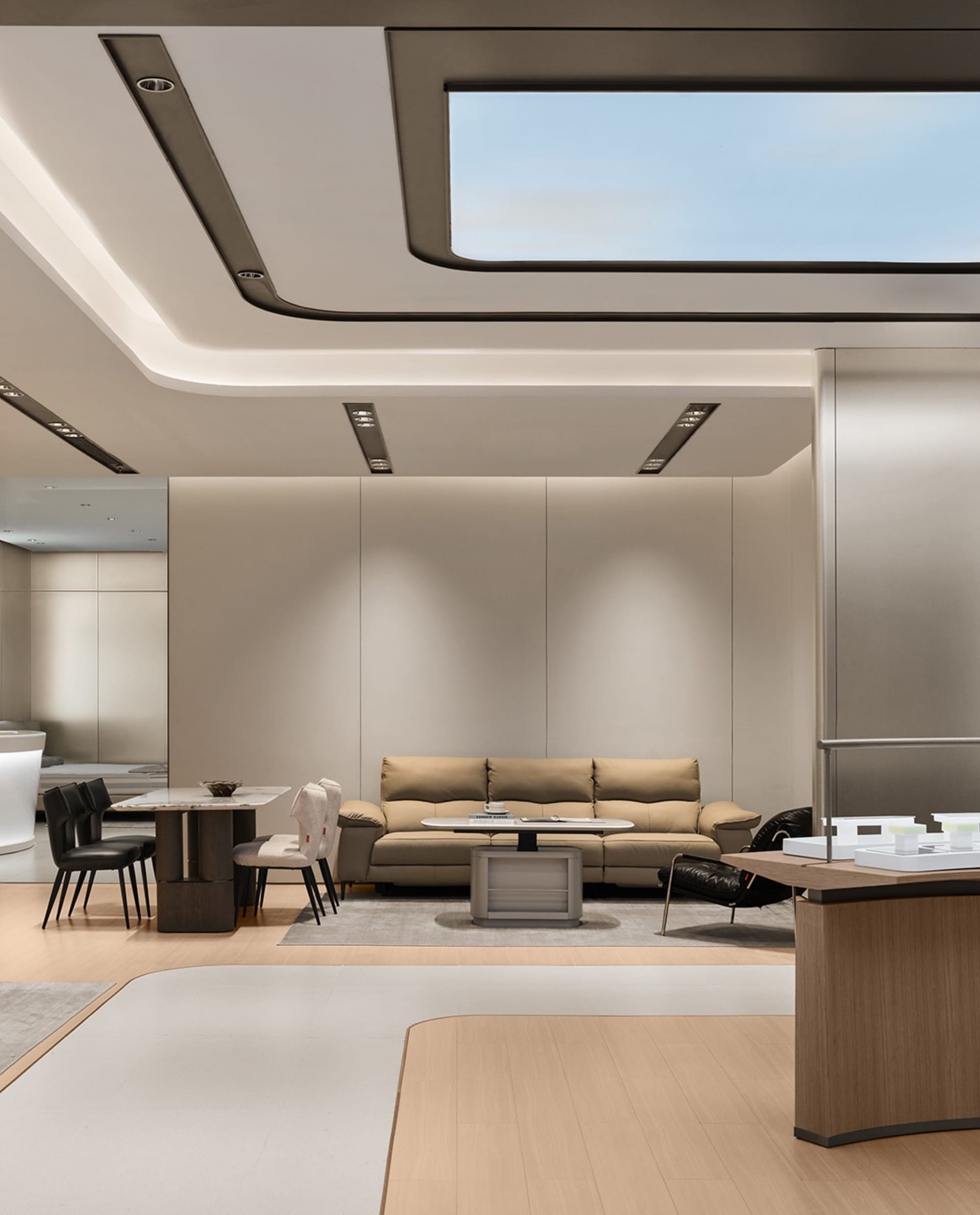 Modern minimalist living room featuring a beige leather sofa, oak wood flooring, marble dining table, black leather chairs, and a large rectangular skylight in a coffered ceiling.
