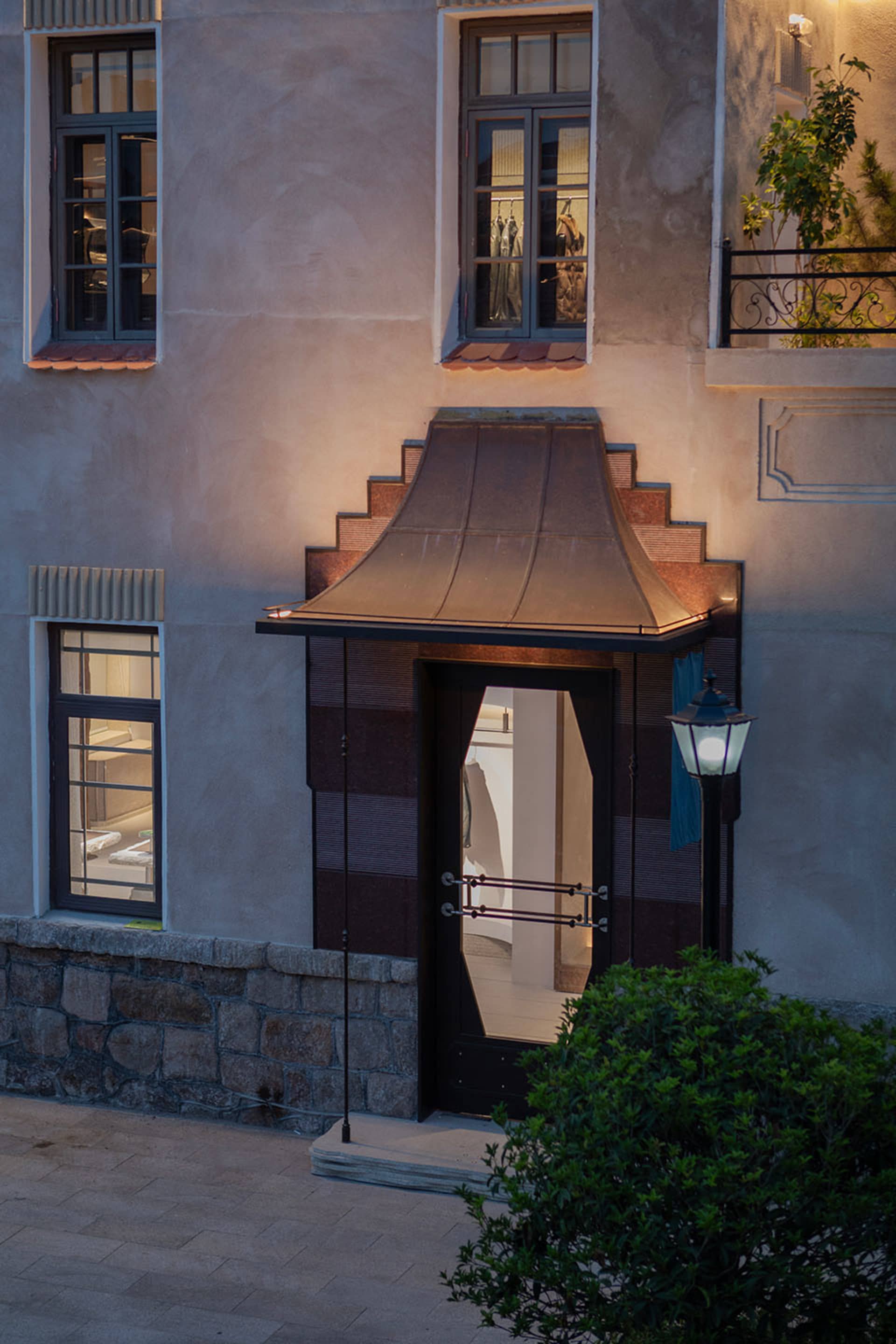 Exterior view of a modern boutique entrance featuring a dramatic stepped Corten steel canopy, dark timber door, and textured stucco facade at dusk.