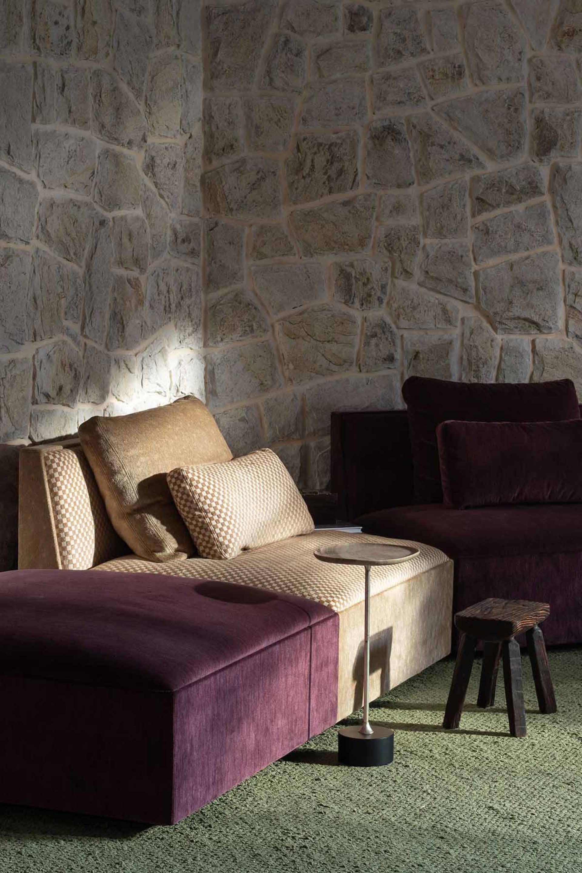 A modern lounge setting featuring a plum velvet chaise and ottoman paired with a beige checkered sofa, set against a textured stone wall with dramatic uplighting, accompanied by a minimalist side table and rustic wooden stool on green carpet.