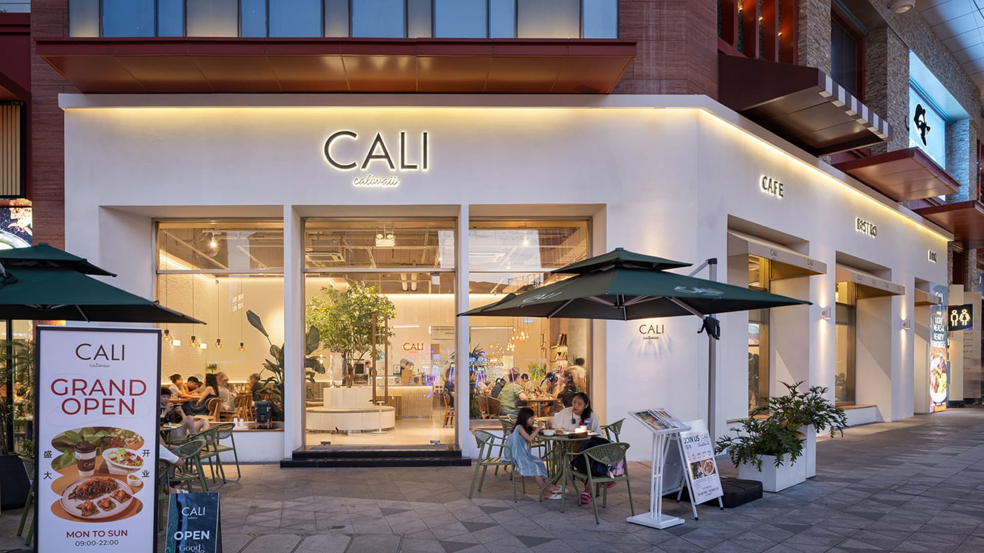 Modern white cafe exterior at night with green outdoor seating, large glass windows, and warm interior lighting showing patrons inside.