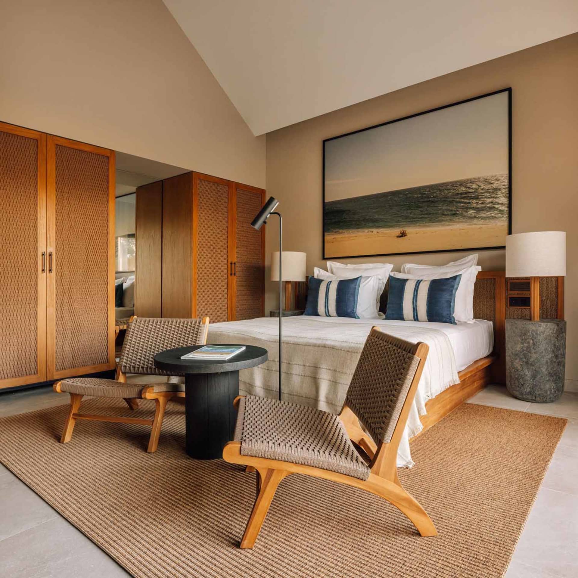 Modern coastal bedroom with light wood teak furniture, woven rattan lounge chairs, neutral linen bedding with blue striped pillows, large beach landscape artwork, and a textured jute rug on tiled floor.