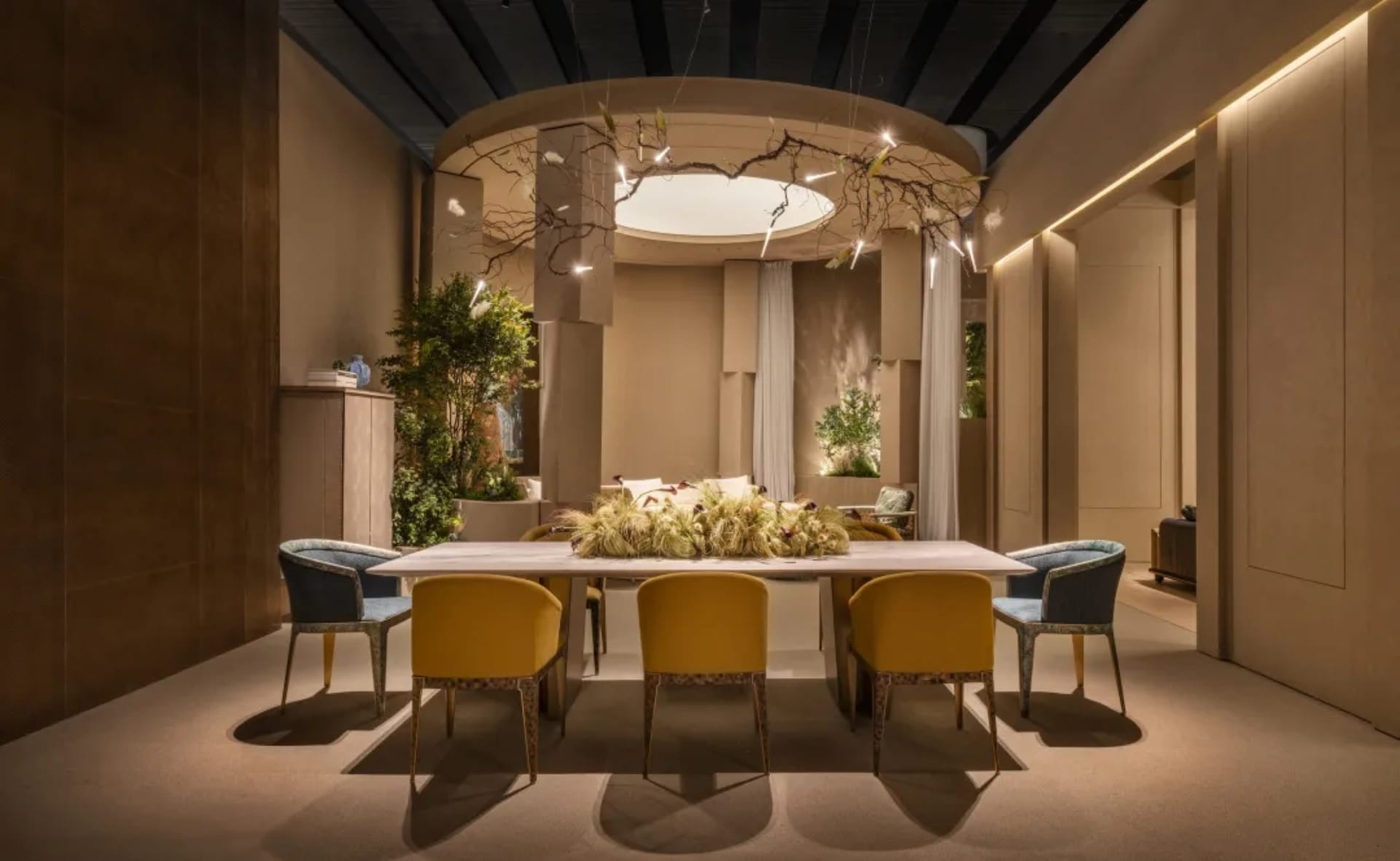 Modern dining room featuring a long rectangular table surrounded by yellow and blue patterned velvet chairs, a large dried floral centerpiece, and a dramatic circular ceiling installation with branching light fixtures.