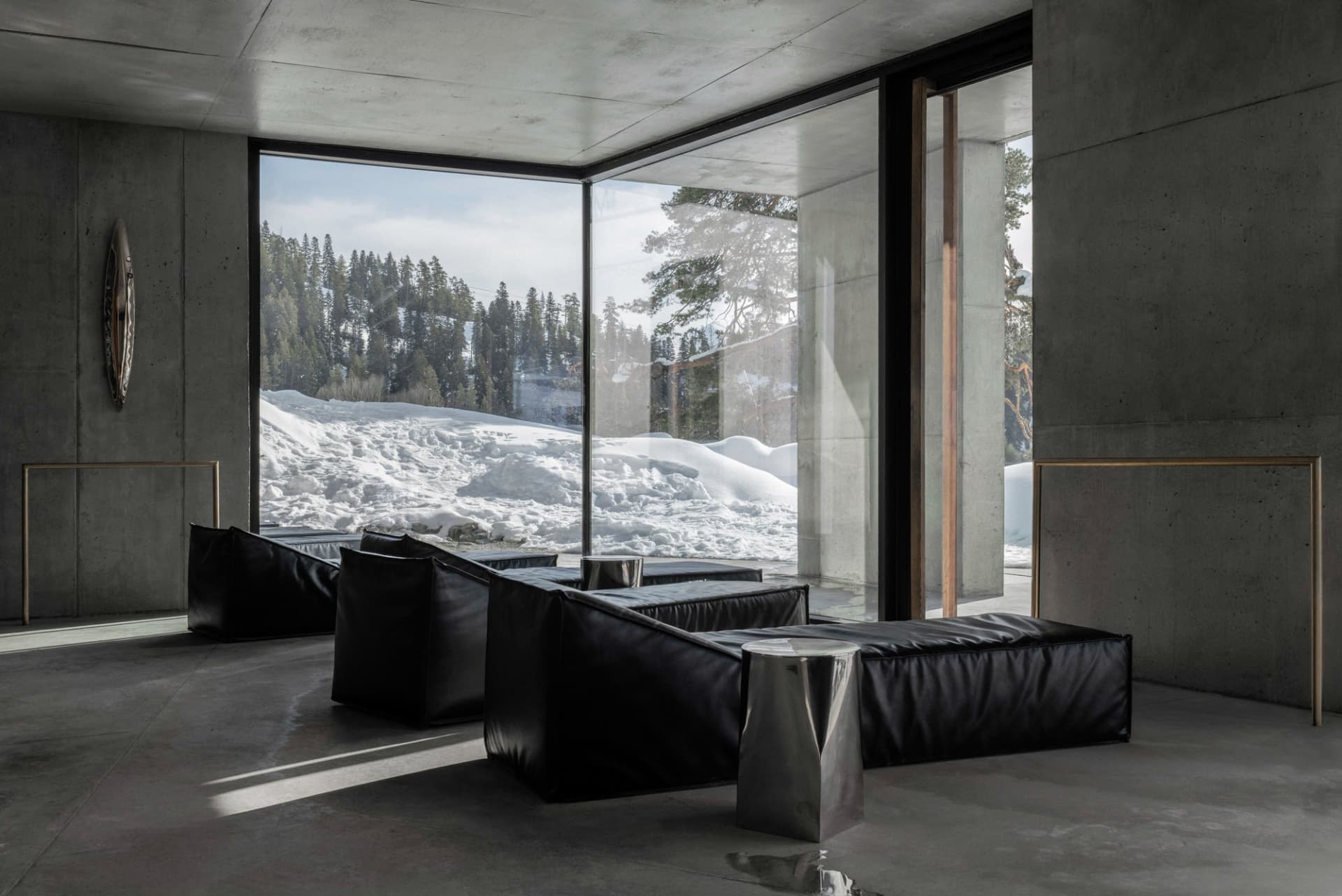 Modern minimalist living room with raw concrete walls, large floor-to-ceiling windows overlooking a snowy mountain forest, black leather modular sofas, and brushed metal side tables.