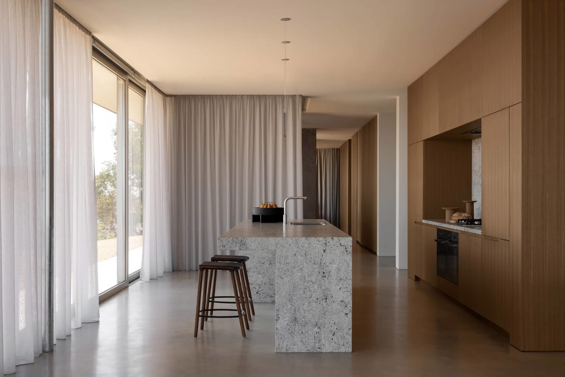 Modern minimalist kitchen with a large grey terrazzo island, wooden bar stools, floor-to-ceiling sheer curtains, and warm oak cabinetry along the right wall.