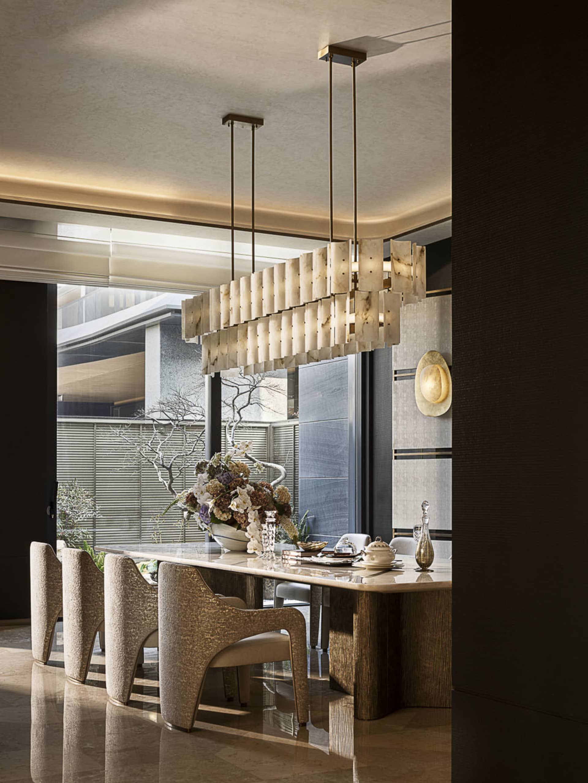 Modern luxury dining room featuring a large rectangular alabaster and brass chandelier, a marble dining table, four textured boucle dining chairs, and a large floral arrangement with a view to an outdoor courtyard.