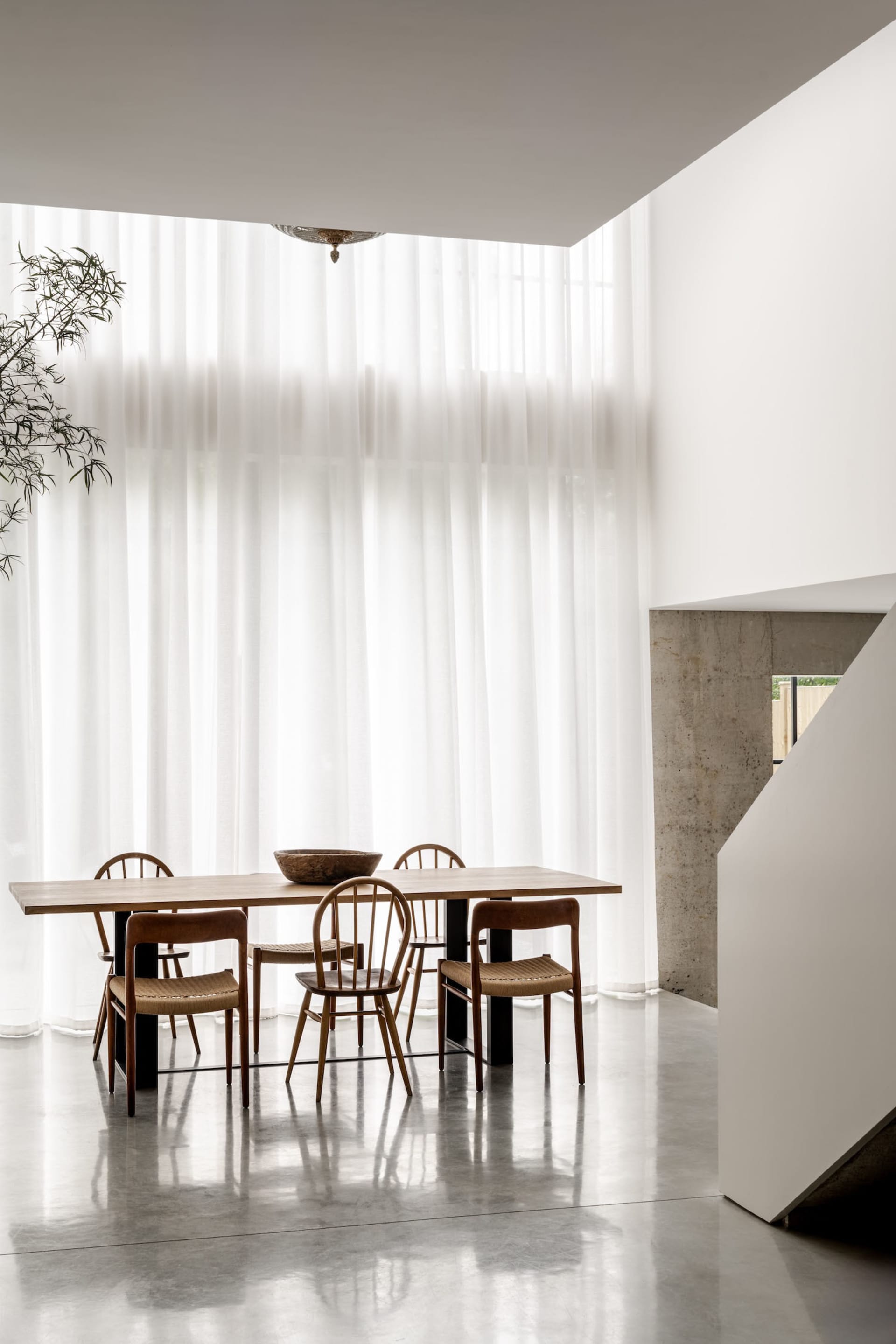 Modern minimalist dining room featuring a light oak rectangular table surrounded by six wooden spindle-back chairs with woven seats, set against a backdrop of floor-to-ceiling sheer white curtains, polished concrete floor, and a raw concrete staircase on the right.
