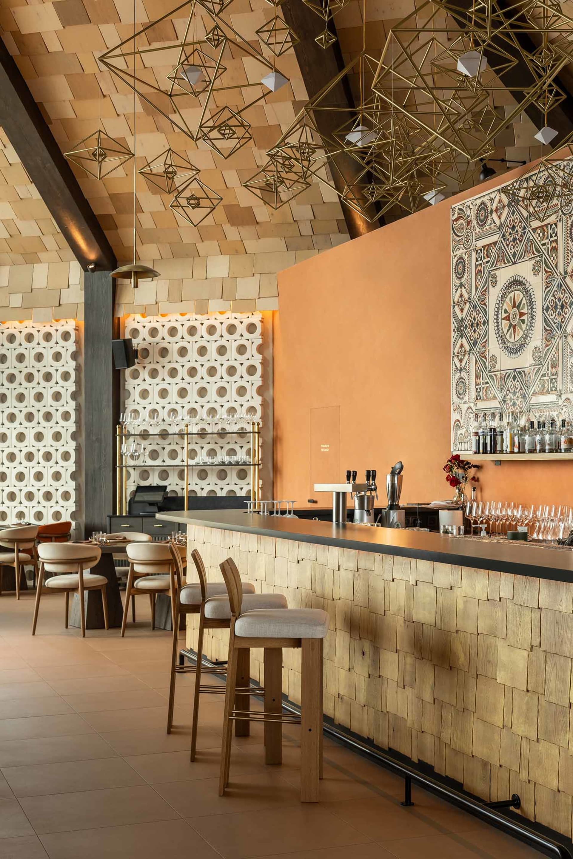 Modern restaurant bar featuring a long counter clad in light wood shingles, wooden bar stools, terracotta accent wall, patterned tile art, and numerous geometric brass pendant lights hanging from a wood-paneled ceiling.